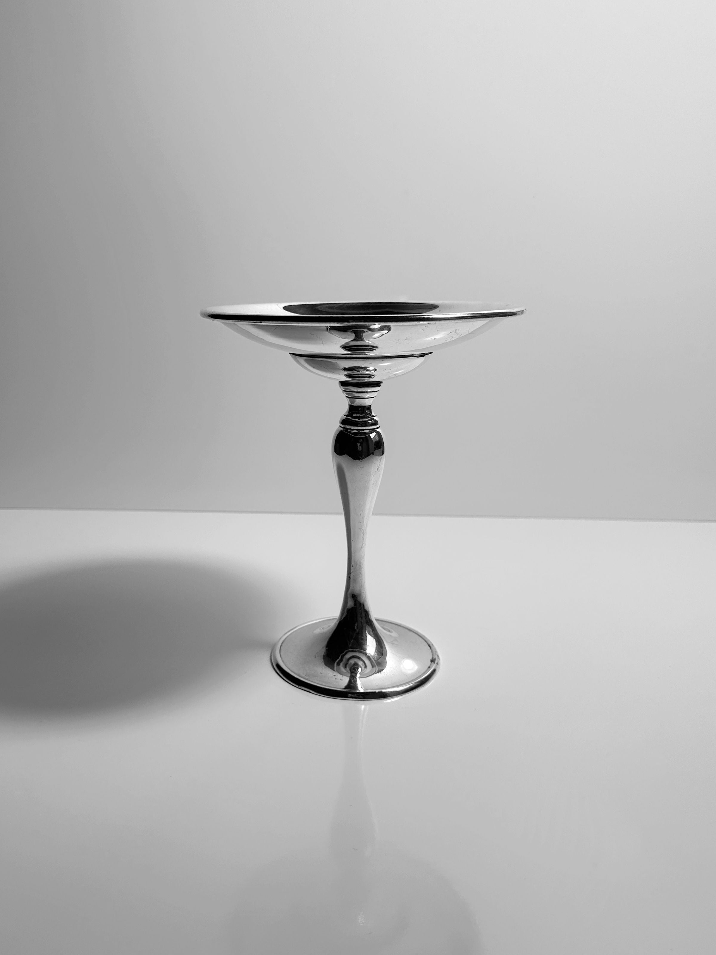 The Dodo Vintage Heavy Silver-Plated Foot Bowl, featuring a wide bowl and slender stem, stands on a glossy white surface against a plain light background.