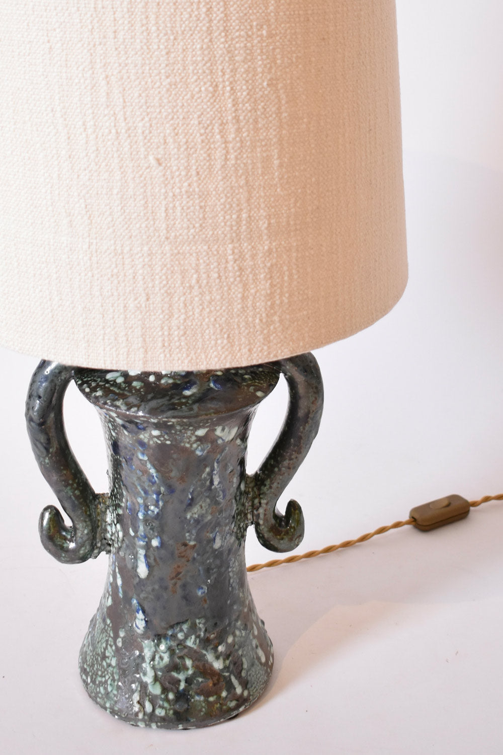 The "Kasaï" Erato Blue and Green Ceramic Table Lamp by Barracuda Interiors features a speckled finish, two curved handles, a large off-white fabric shade, and a twisted brown cord—an eye-catching accent for any interior.