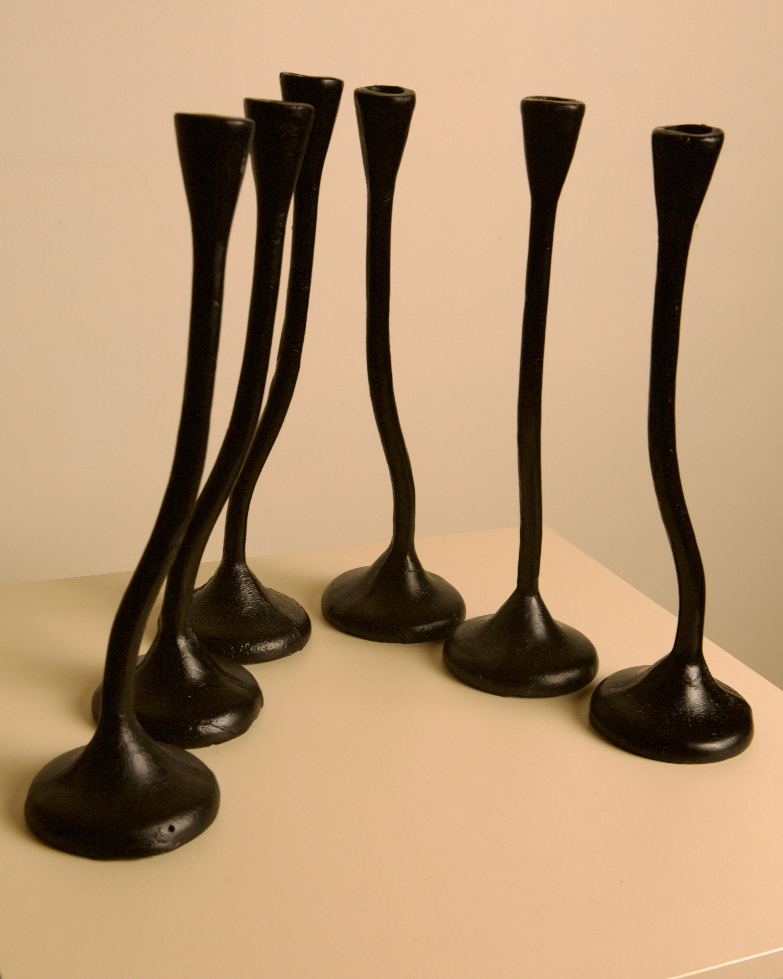 The Set of 5 "Twisted" Candleholders by Treaptyque feature black cast aluminum with wide bases and narrow, curved stems inspired by 1980s sculptural design, arranged on a beige surface against a light background.