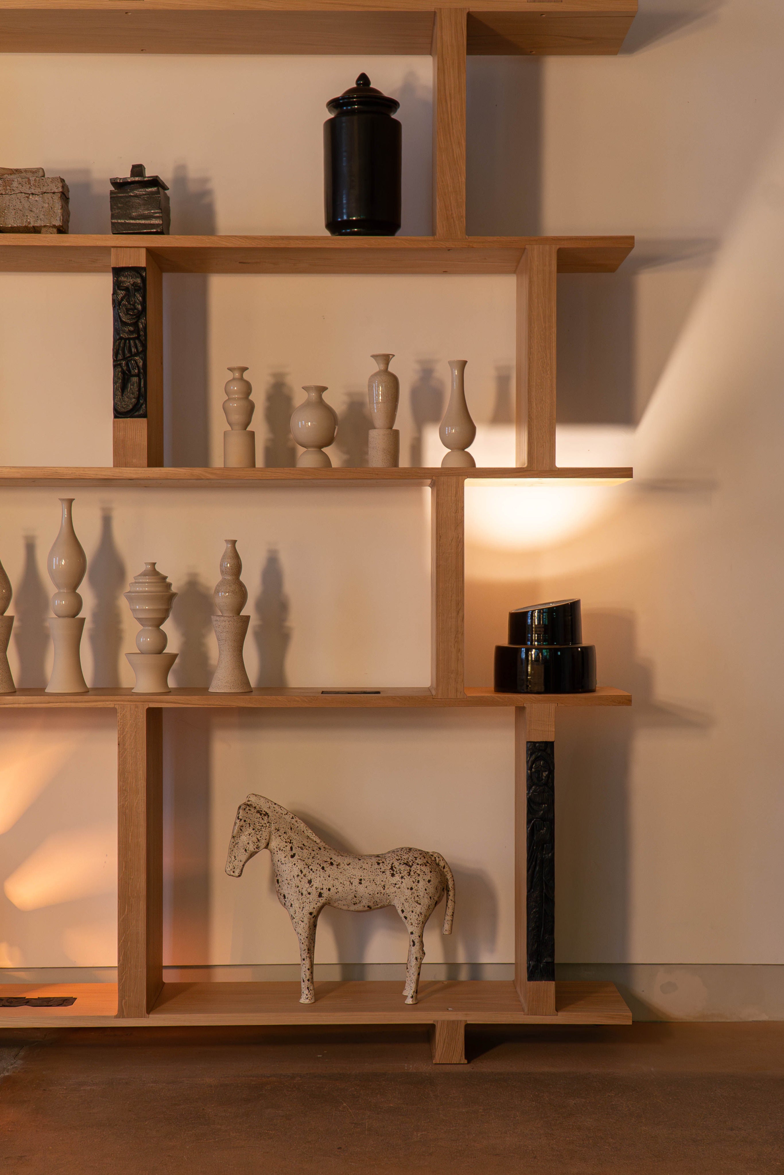 A wooden shelf displaying hand-thrown ceramic vases, jars, and a horse figurine is softly illuminated by COSEINCORSO's The Pillar - Minimalist Table Lamp in Ceramic, casting warm shadows on the beige wall behind.