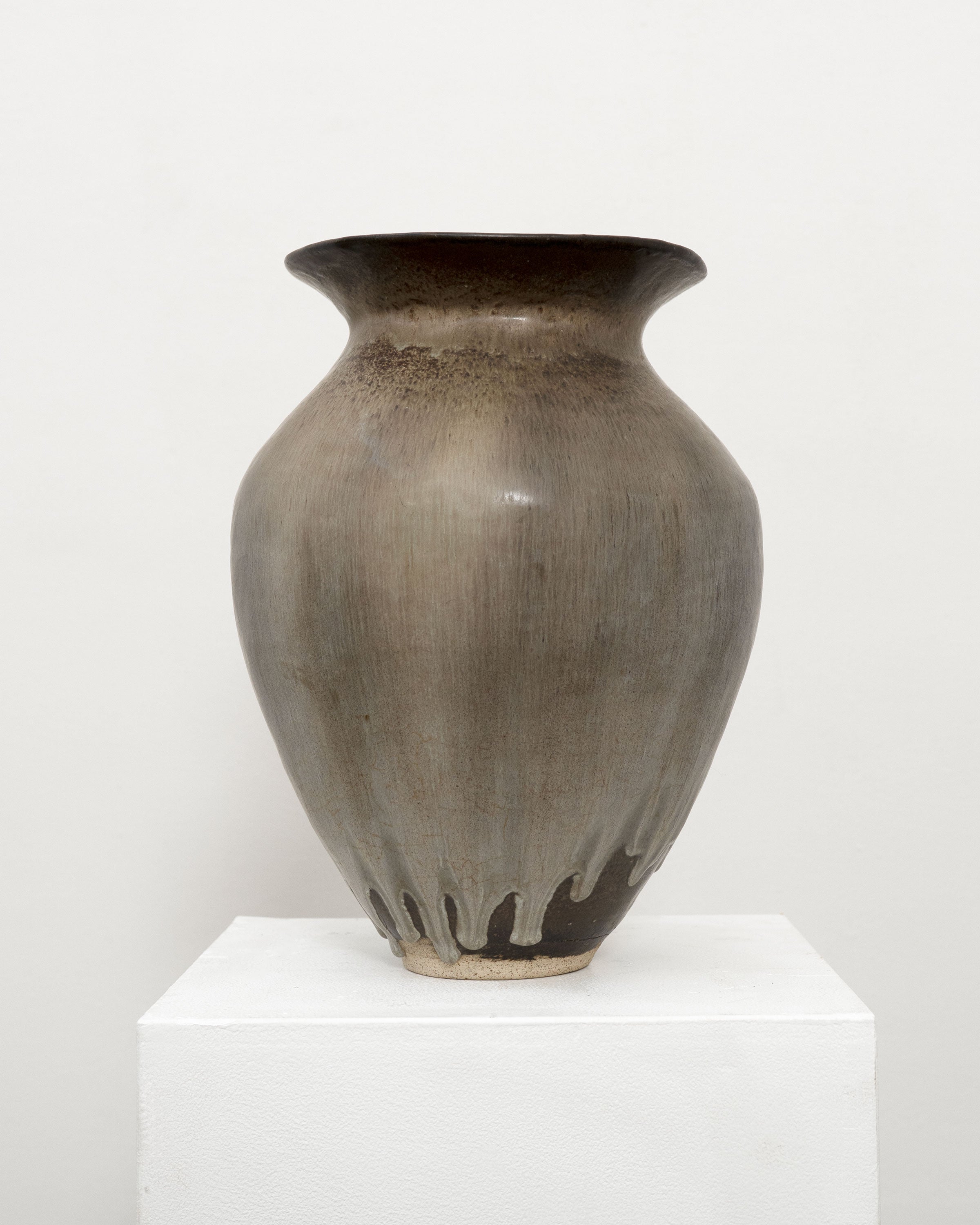The Tom Bogle Hand Built Studio Pottery Vase is a tall, wide stoneware piece with a flared rim, brownish-grey glaze, and white drips at the base. It rests on a white pedestal against a plain white background.