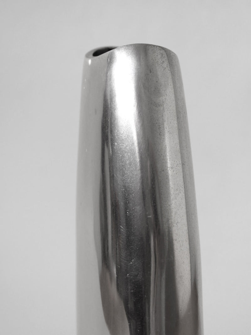 The Vintage Stories Polished Aluminum Flat Tall Vase, inspired by 1960s design and made in Germany, features a smooth, reflective curved surface and narrow opening, displayed against a plain light gray background.