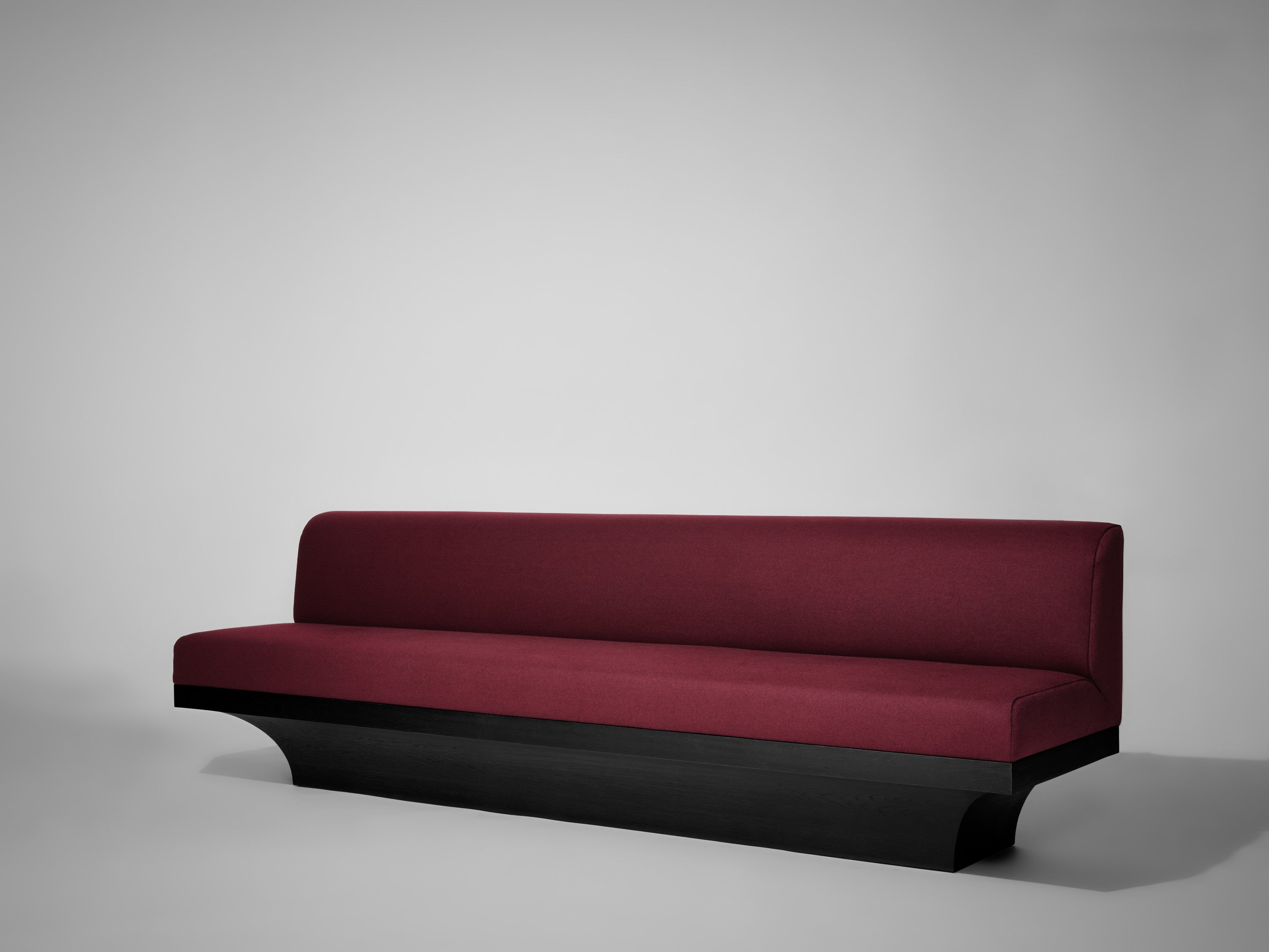 The Mokko Acer Sofa is a minimalist maroon upholstered bench with a black wooden base, ideal as dining room seating or a living room accent, shown against a plain light gray background.