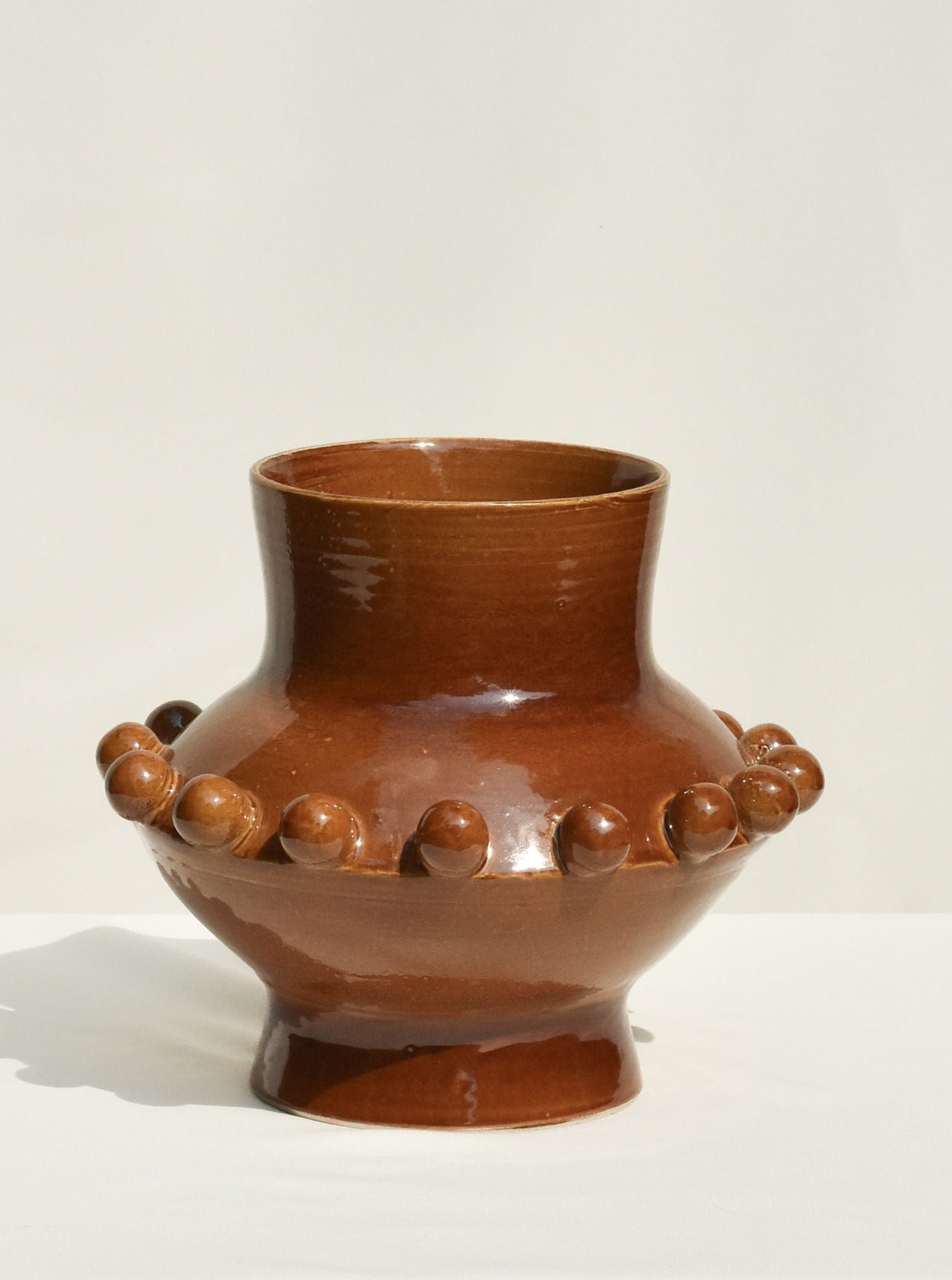 The UFO Vase in Amber by Landy Rakoto Ceramics features a glossy brown finish, wide base, narrow neck, and rounded protrusions—a striking 70s-style decor piece set against a light background.