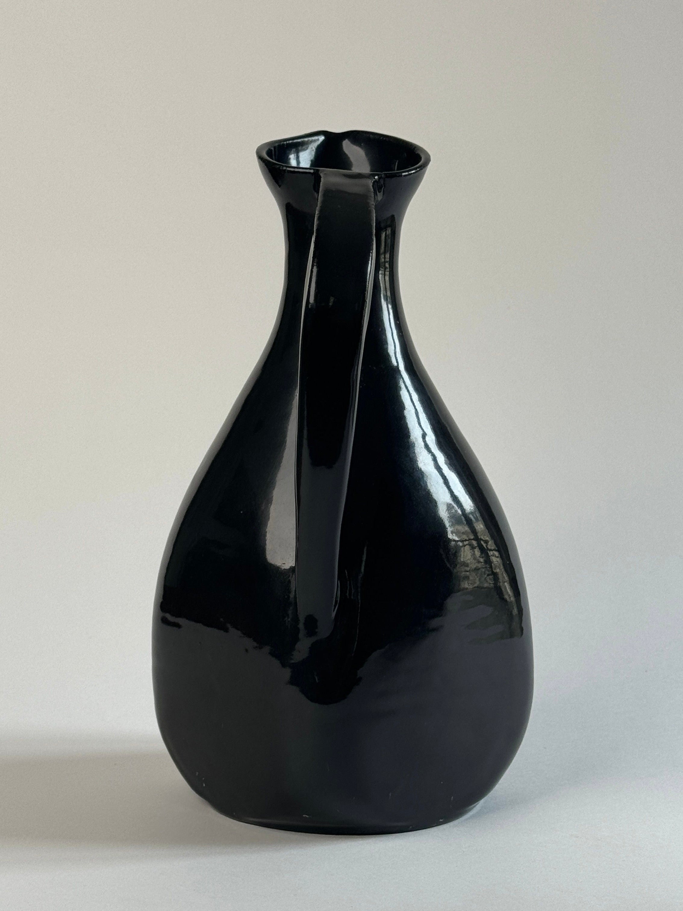 A pair of glossy black Accolay Ceramic Carafes by Figuresdesign, featuring wide bases and narrow necks, shine against a light gray background as their smooth surfaces reflect the light.