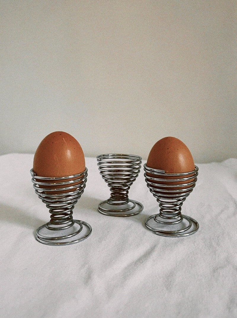 Three wire spiral egg cups with brown eggs on white linen surface