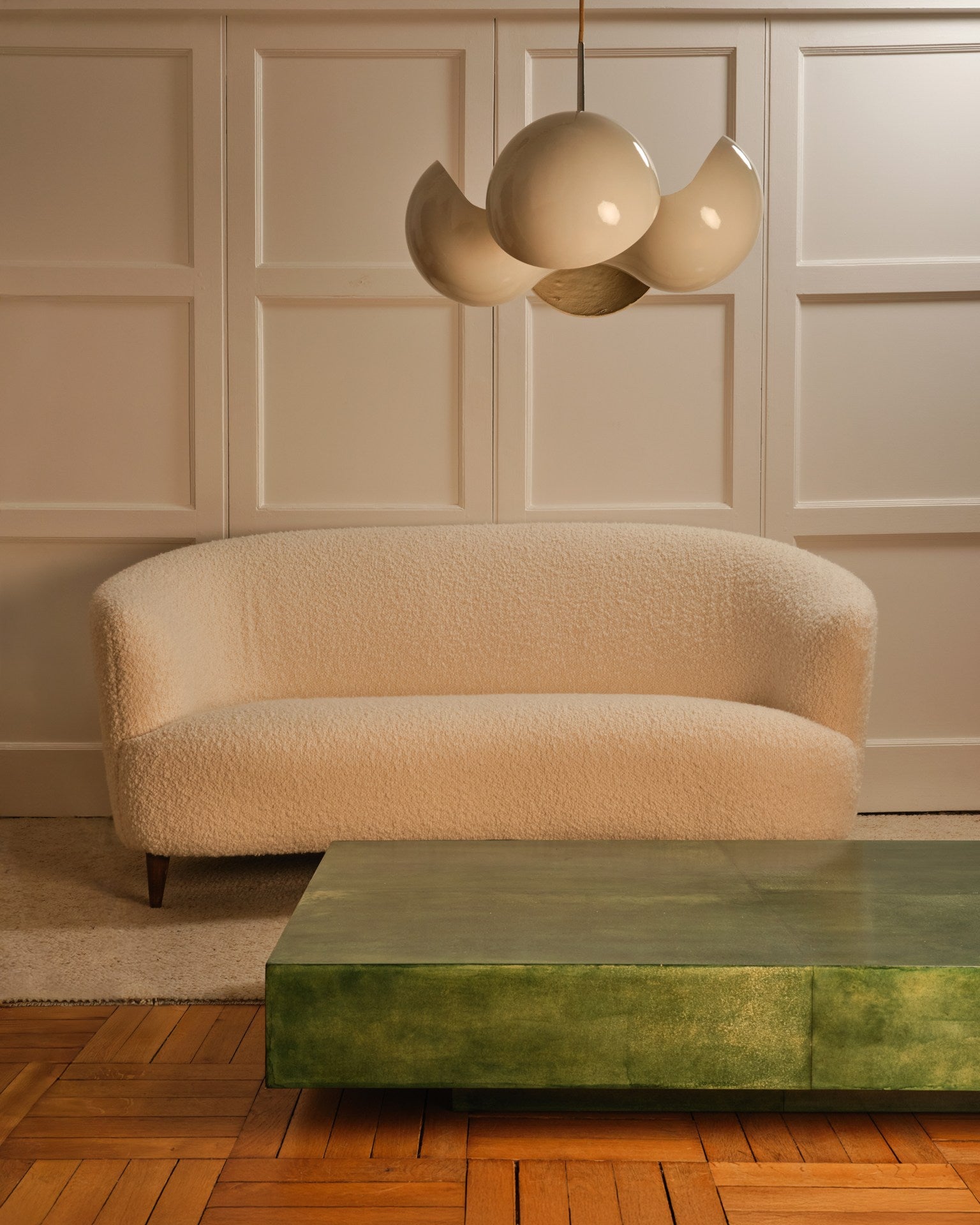 A minimalist living room with a textured white loveseat, Galerie Vauvart's green parchment table as the coffee table, wood-paneled flooring, a sculptural white pendant light, all set against white paneled walls.