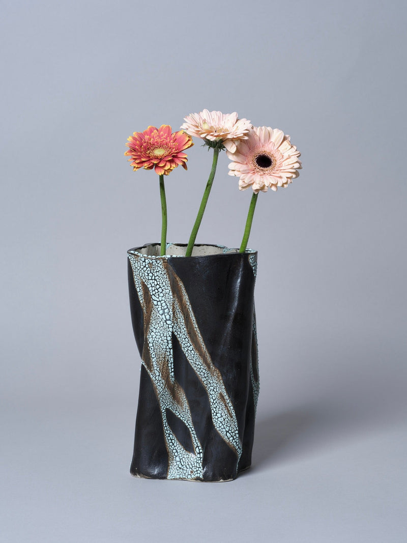 Three gerbera daisies—one orange, two pale pink—are arranged in the Swirly Vase Brown and Light Blue by Astrid Öhman, set against a plain gray background.