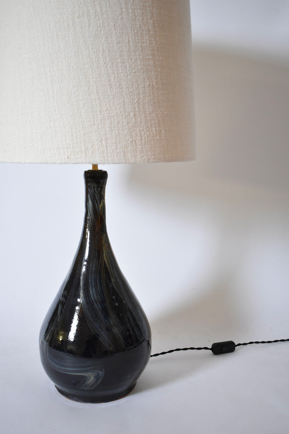 The Black Ivory Brush Lamp by Barracuda Interiors features a glossy, swirl-patterned teardrop ceramic base in dark tones and a large off-white textured fabric shade. Includes a black cord with inline switch.