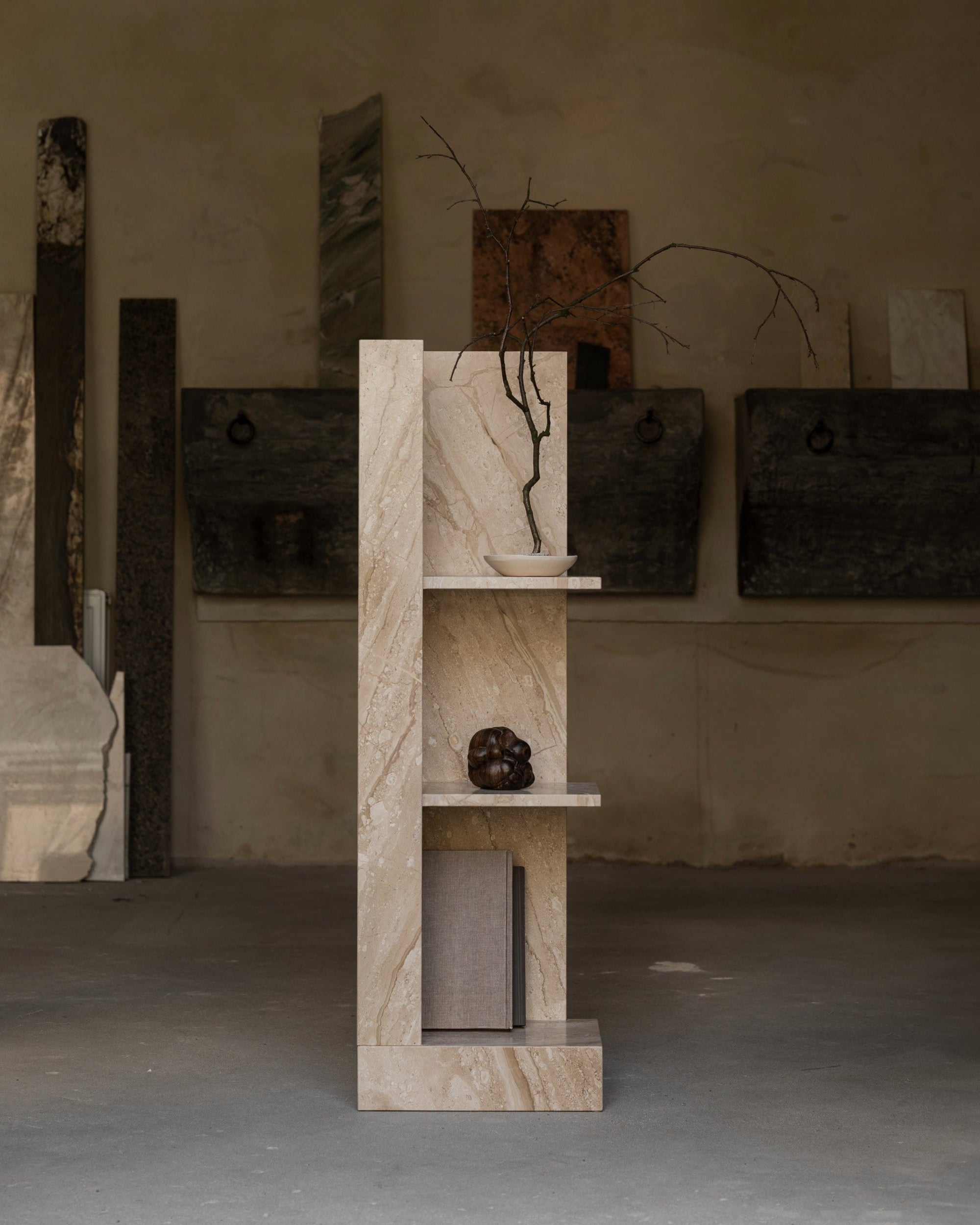 The un'common CONE Marble Shelf features three minimalist shelves displaying a bowl with a branch, a dark accent piece, and a closed book. Stone slabs rest against a textured beige wall, highlighting this space-saving design.
