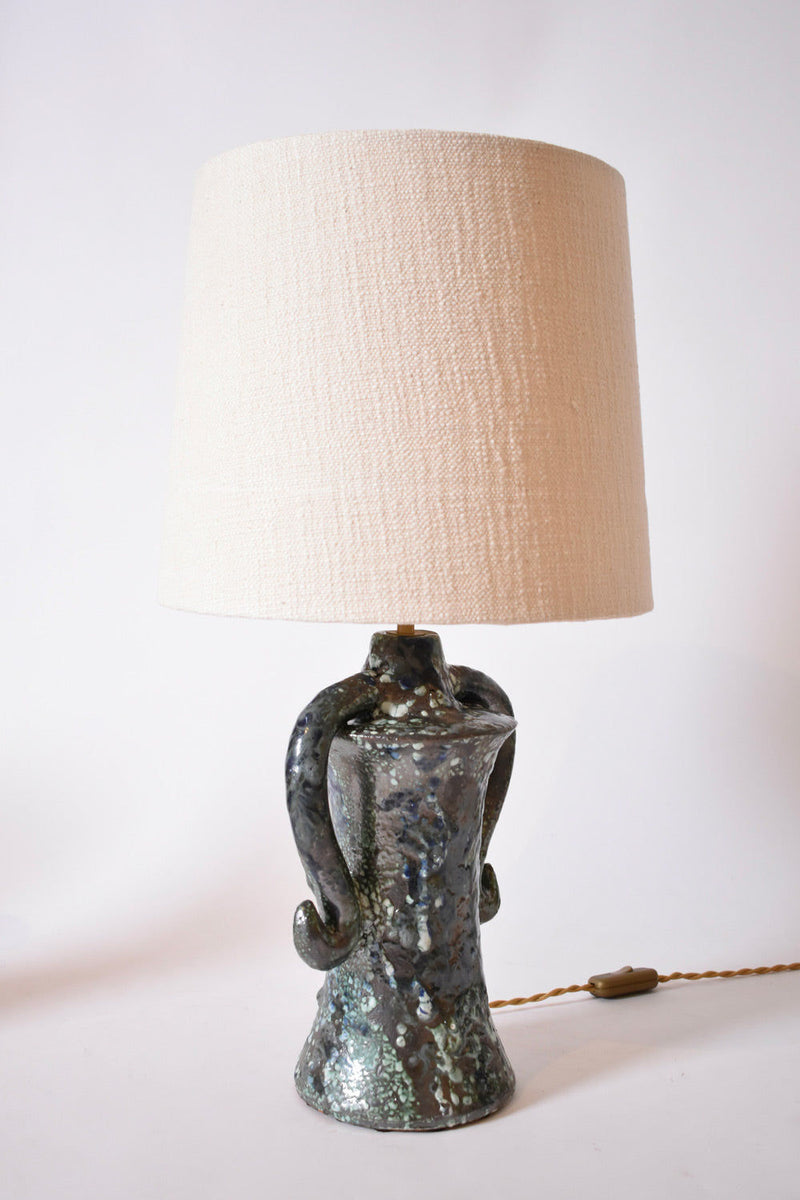 The "Kasaï" Erato Blue and Green Ceramic Table Lamp by Barracuda Interiors features a sculpted, textured base with two curved handles and an off-white fabric shade. The cord and switch are visible, making it a statement piece for any space.