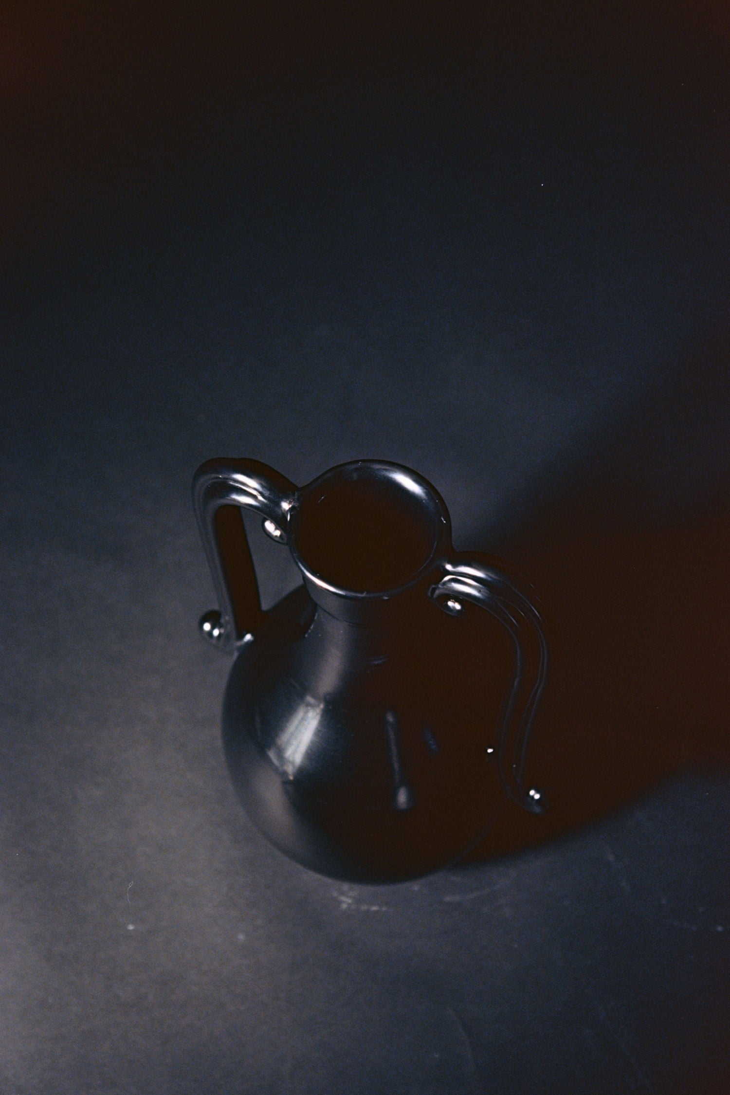 The Fiasca - Minimalist Vase in Ceramic by COSEINCORSO features a matte black finish and dual curving handles. Handmade in Italy, it rests on a dark surface under soft light, highlighting its refined artisanal craftsmanship.