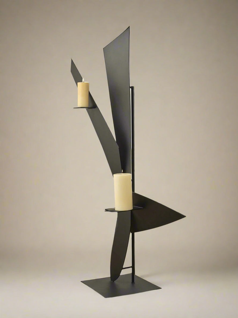 Animate Objects’ Bird Candleholder features a modern, abstract black powder coat design with geometric shapes and two beige pillar candles on separate platforms, showcased against a plain beige background.