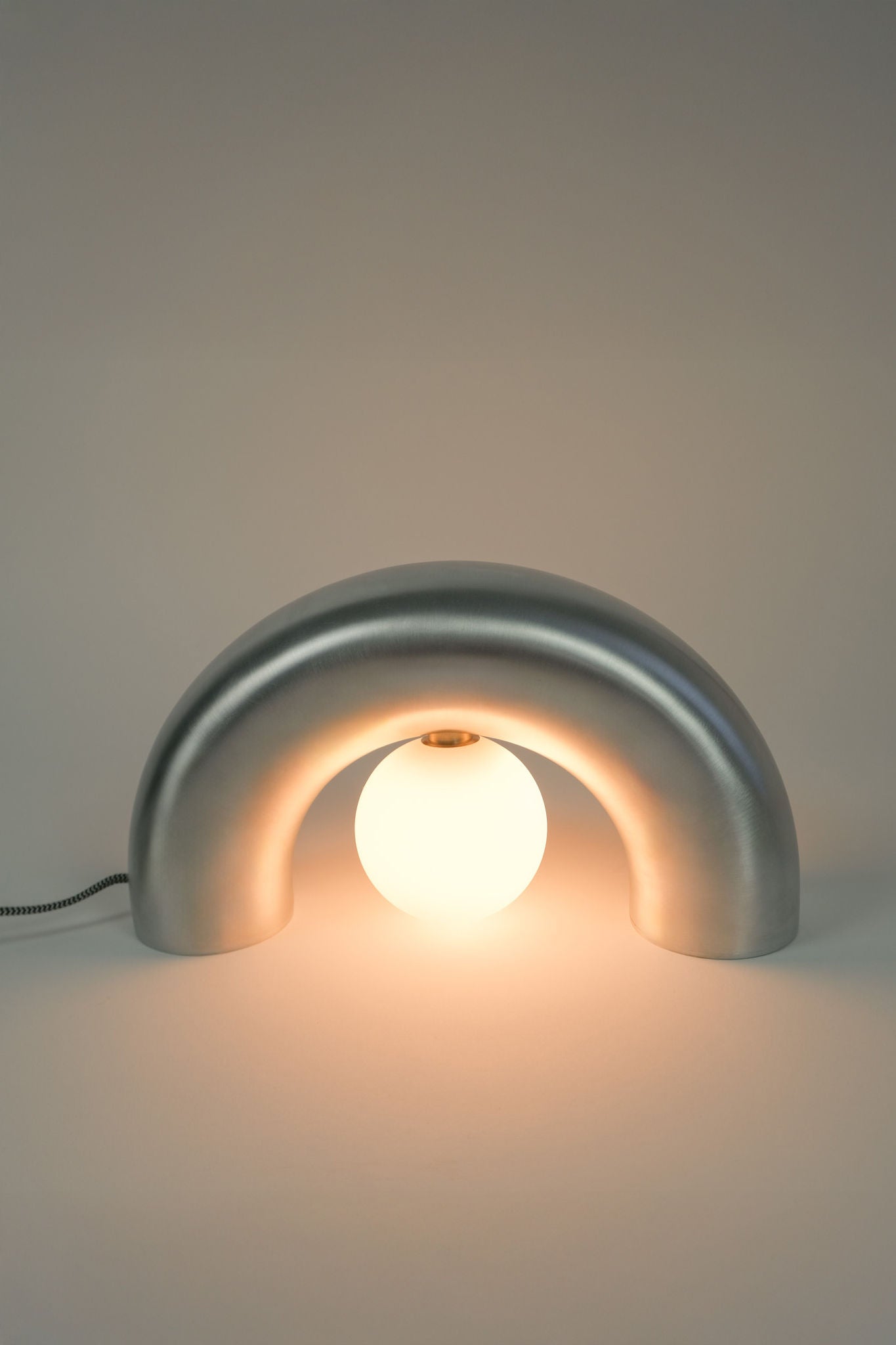 The Arcora Underglow Aluminum Arc Lamp by Heako Studio features a sleek metallic arched base and central glowing bulb, casting soft light for a calming effect against any neutral background.