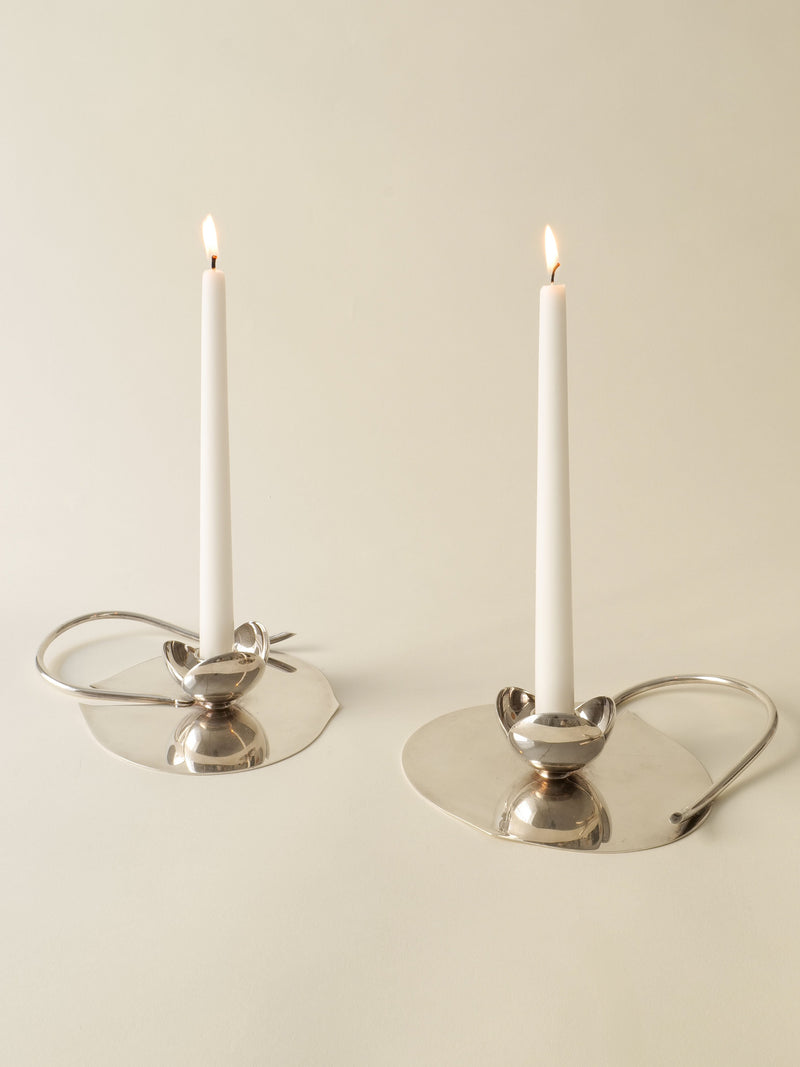 Two lit white candles in sleek, modern Candle Holder Lino Sabattini 1970s by Collection apart, featuring silver-plated wide flat bases and curved handles, placed on a neutral surface against a light beige background.
