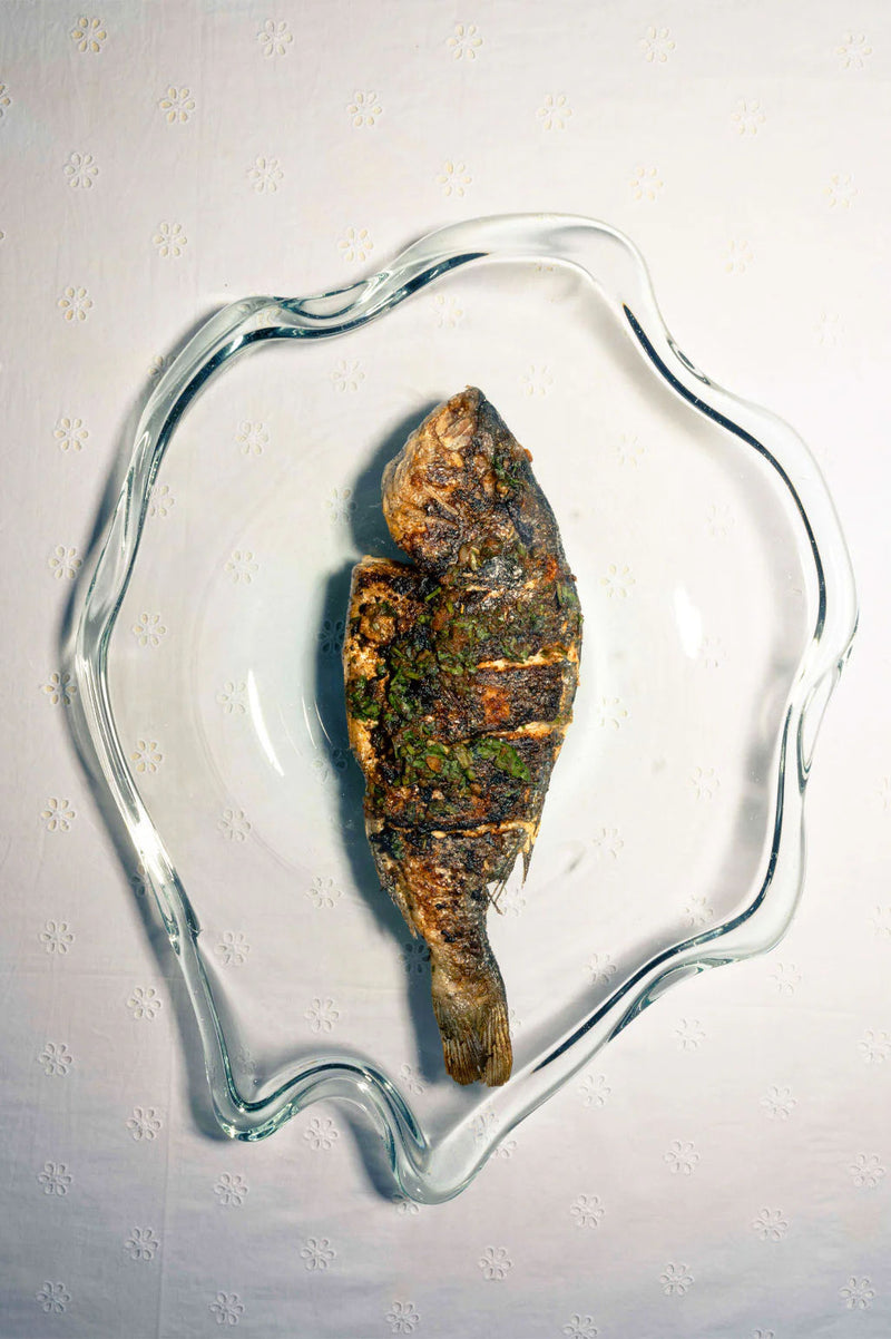 A whole grilled fish, seasoned with herbs, is served on the Completedworks Pond Recycled Glass Serving Plate in Clear, its wavy edges displayed atop a white patterned tablecloth.