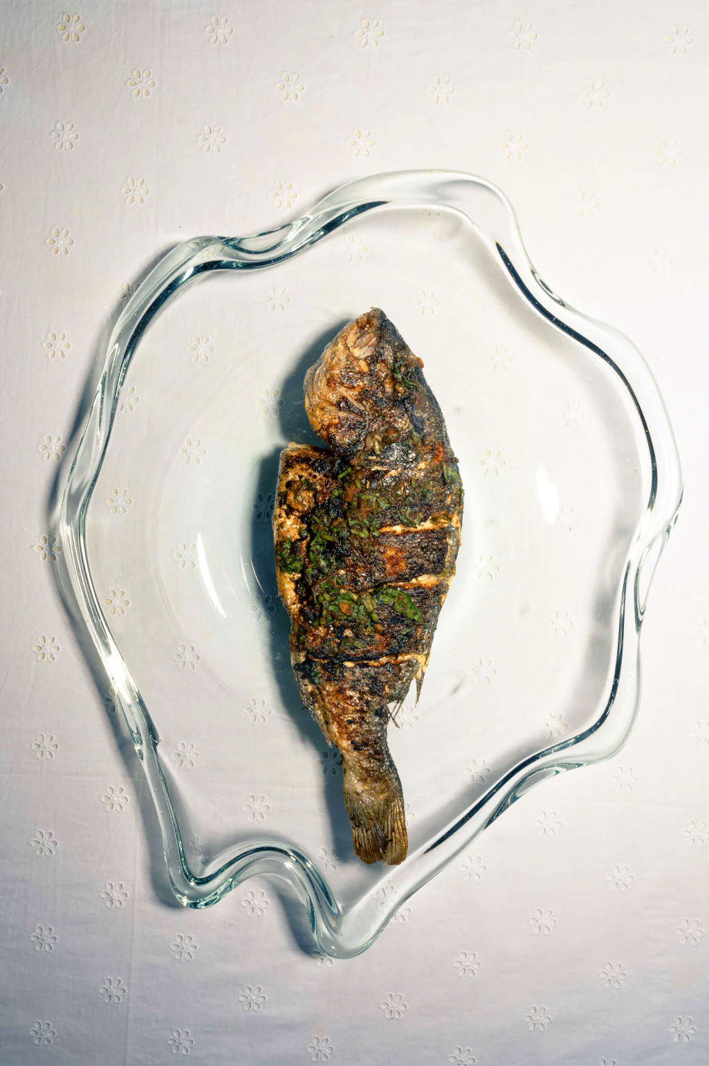 A whole grilled fish, seasoned with herbs, is served on the Completedworks Pond Recycled Glass Serving Plate in Clear, its wavy edges displayed atop a white patterned tablecloth.