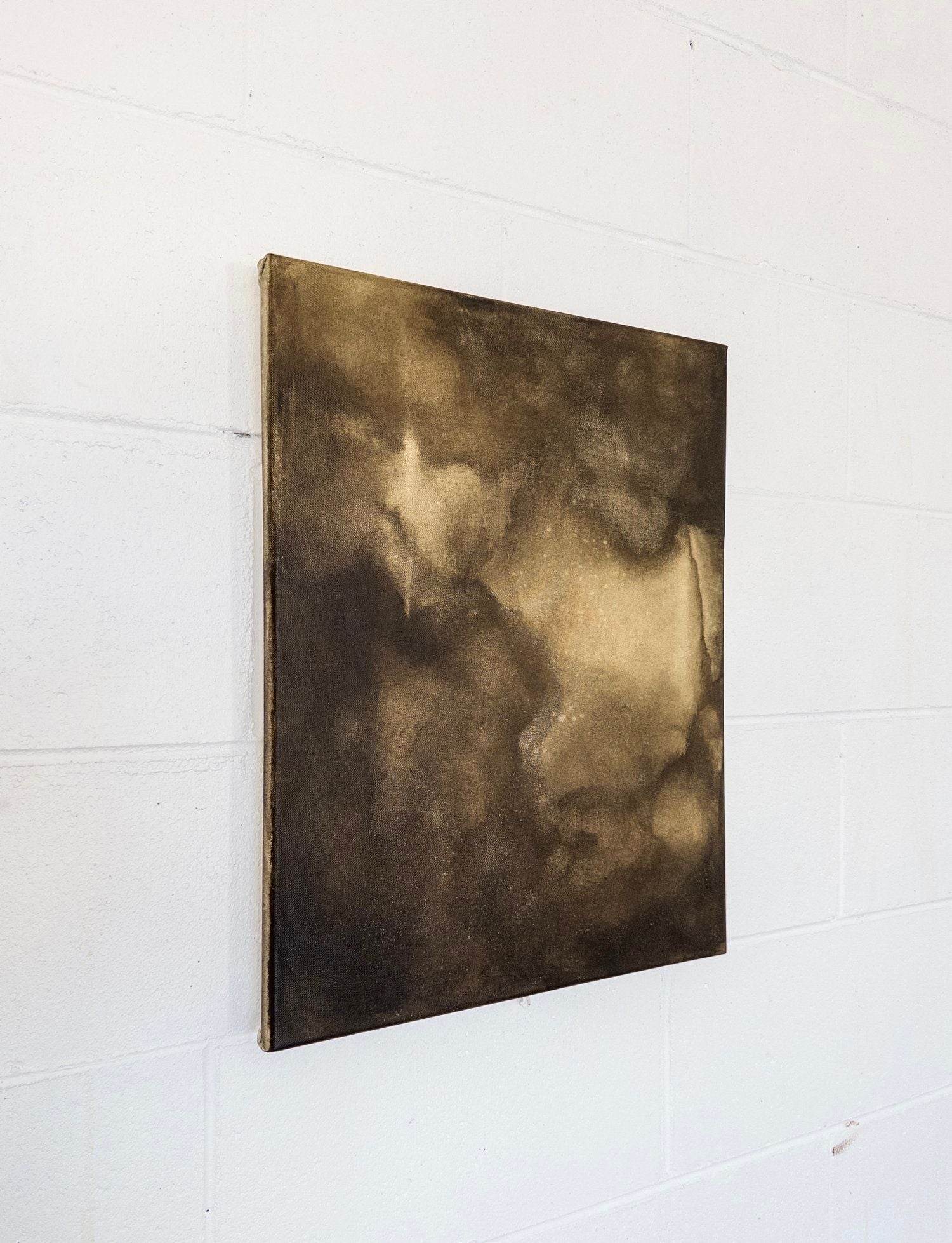 “Midnight Hours Burn Brightest” by Natasha France, a square abstract original in dark brown and gold mixed media, features soft, cloudy patterns without distinct shapes and is shown hanging on a white brick wall.