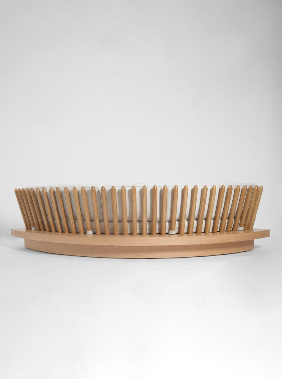 Featuring an oval base and outward-fanning vertical slats, this modern wooden bench from Barracuda Interiors echoes the elegance of the San Romano Round Oak Sofa, offering a striking open circular design against a plain background.