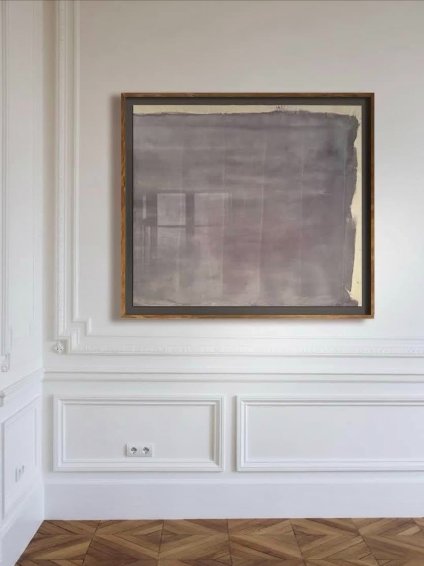 A minimalist room with white paneled walls and chevron wood floors features Médecine’s "Gray Mauve" Pigments, Ink on Raw Canvas in a wooden frame, hanging above an electrical outlet.