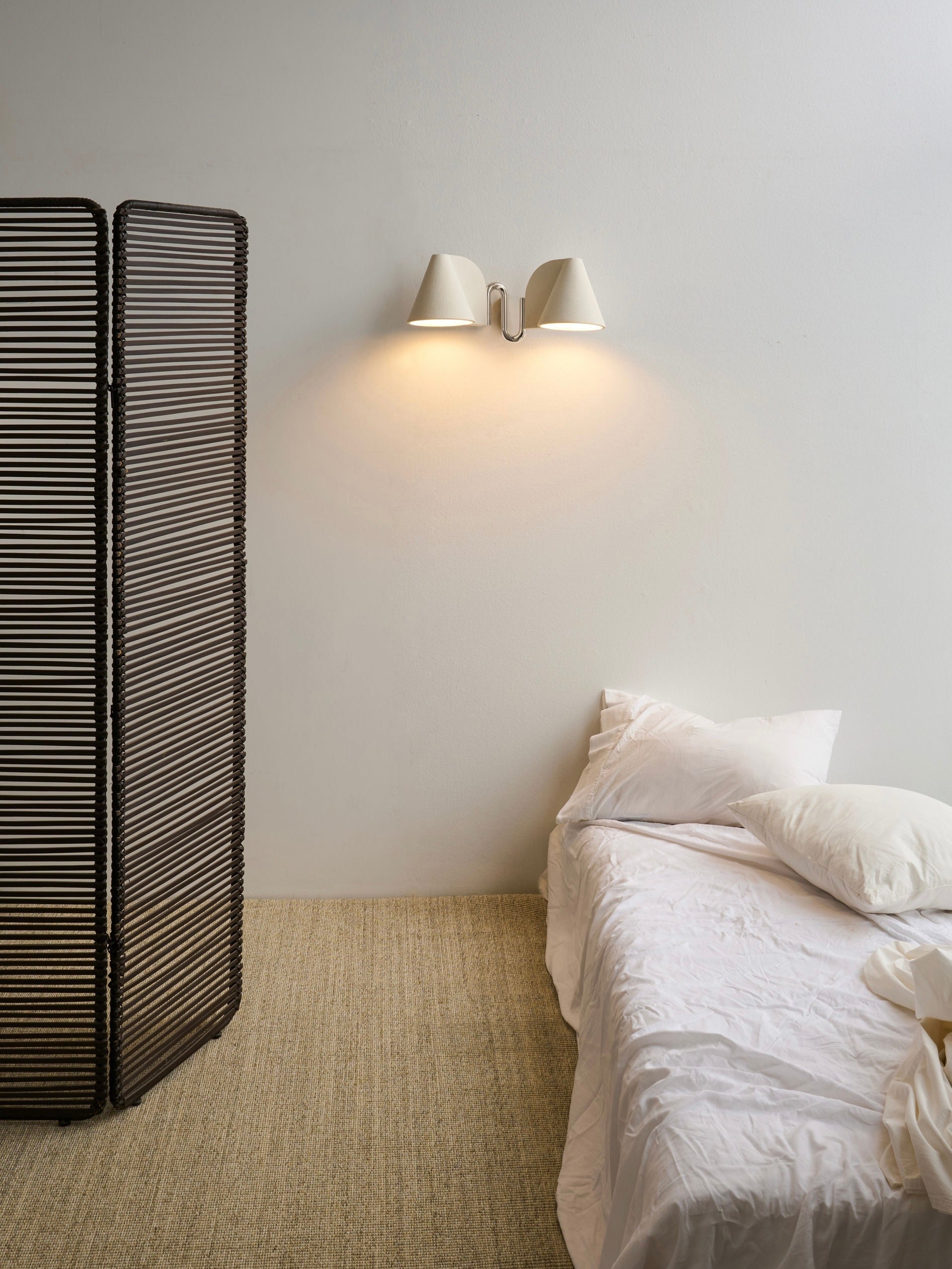 A minimal bedroom with a bed in white linens, a textured beige carpet, a black woven room divider, and the 'Synapse' Wall Light by Mariza Galani casting warm illumination on a plain white wall.
