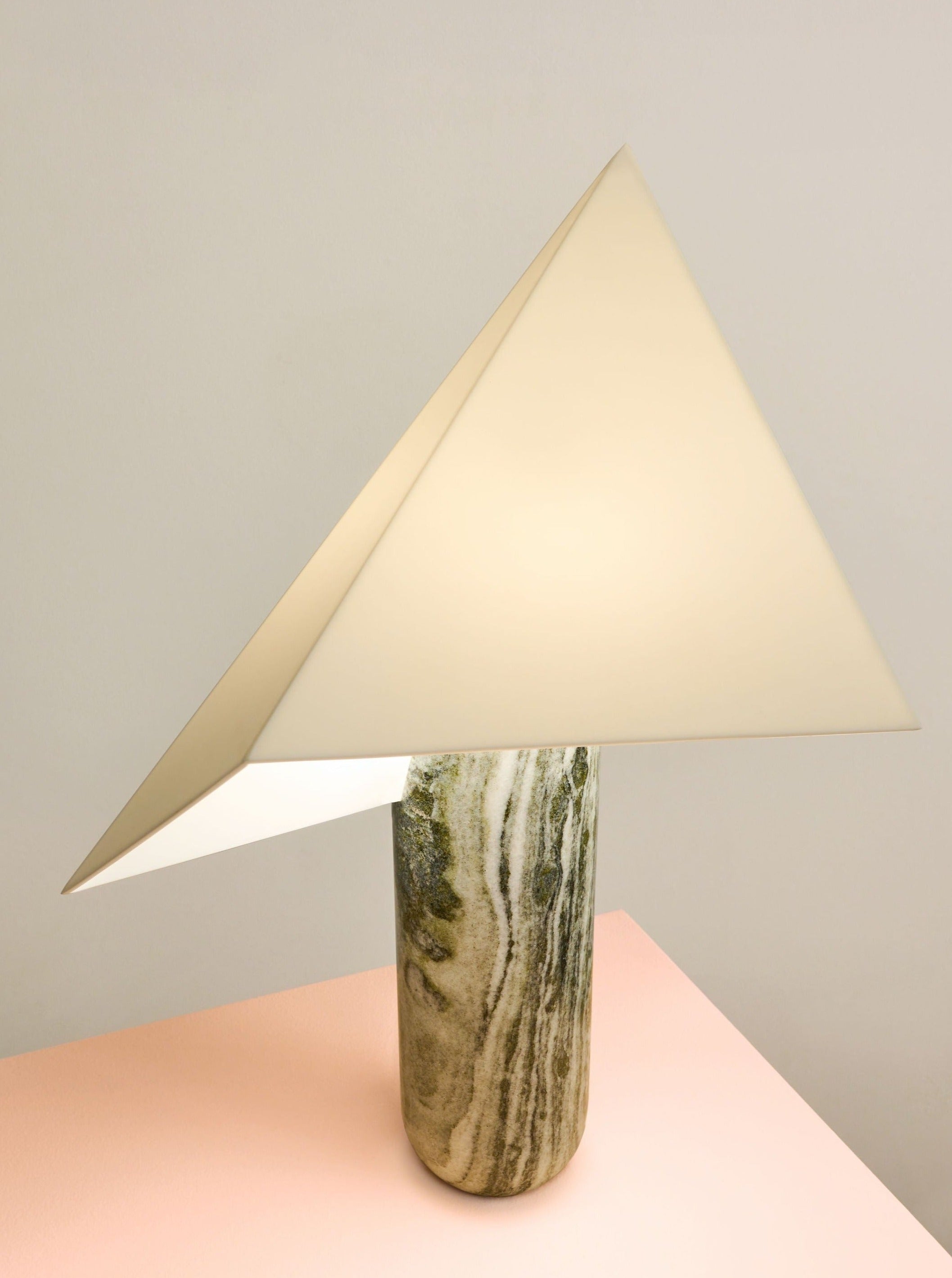 The Acme - Greek Marble table light by Mariza Galani features a triangular cream shade and handcrafted Greek marble cylindrical base, displayed on a light pink surface against a plain background.