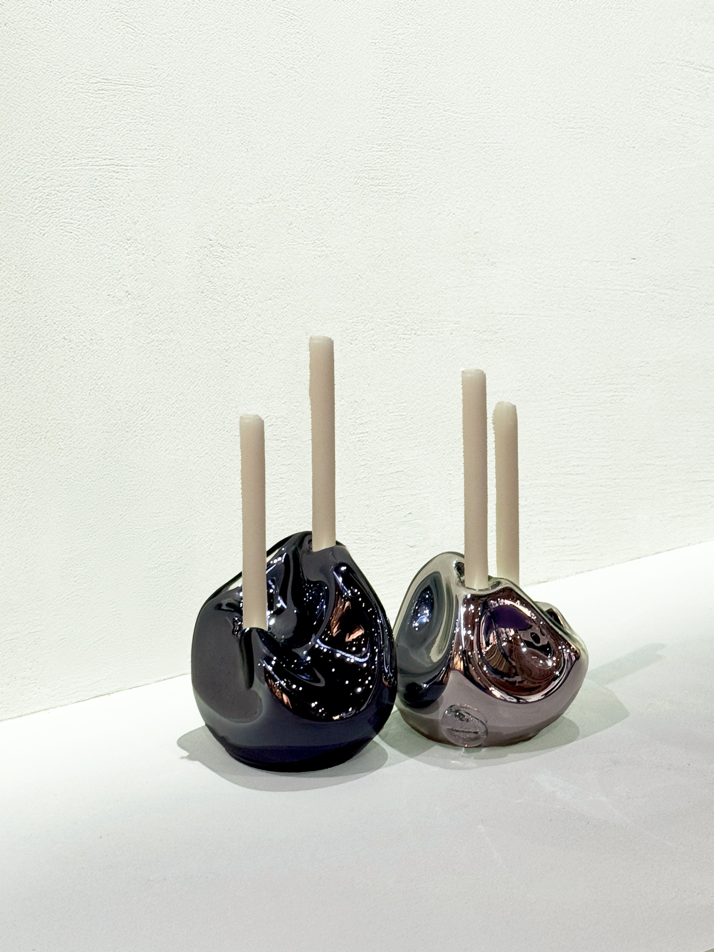 The HOMA Lumi Candle Holder V, in black and metallic silver, each display two unlit white candles on a white surface with a textured backdrop, their sculptural forms echoing each other like molten glass in motion.