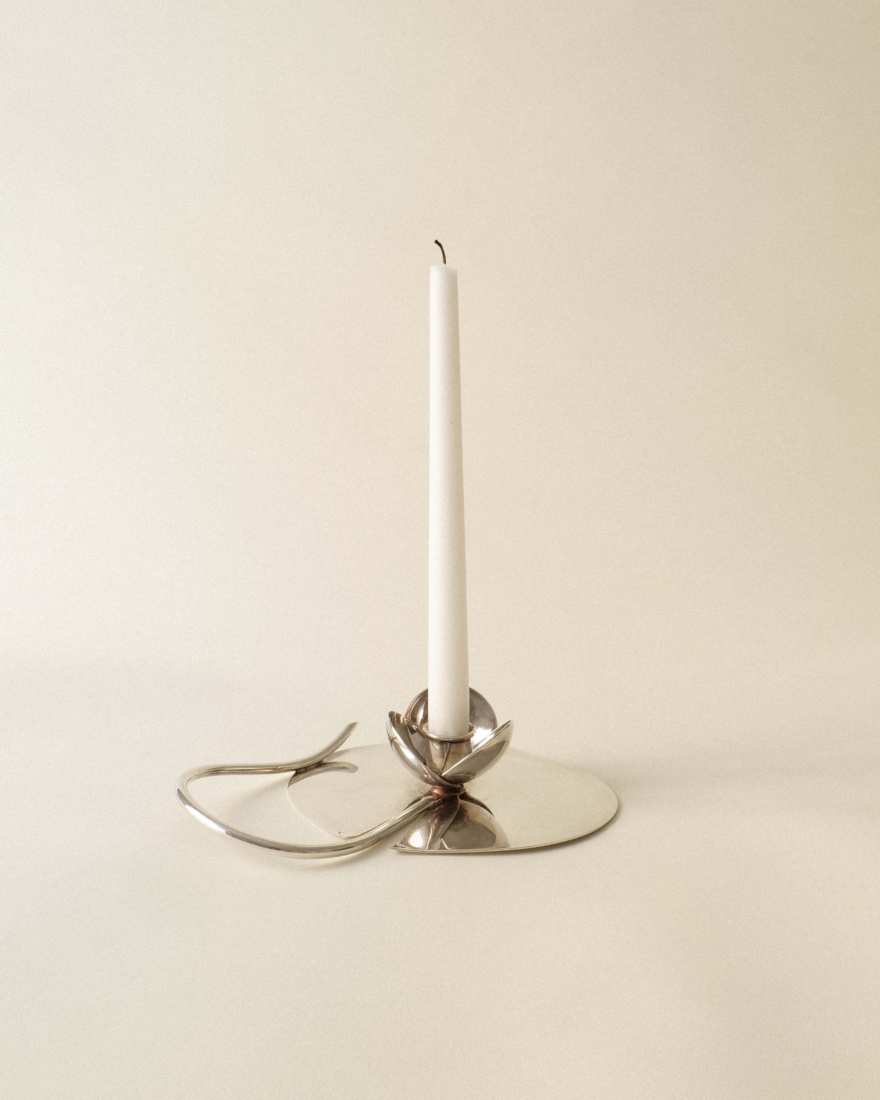 A white taper candle is displayed in the Candle Holder Lino Sabattini 1970s by Collection apart, featuring looping, abstract silver-plated shapes, set against a plain beige background.