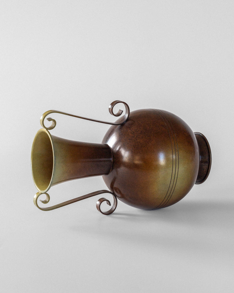 The Spigel Swedish Art Deco Bronze Vase, featuring a round body, narrow neck, and ornate spiral handles, is shown lying on its side against a plain white background, highlighting its elegant design.
