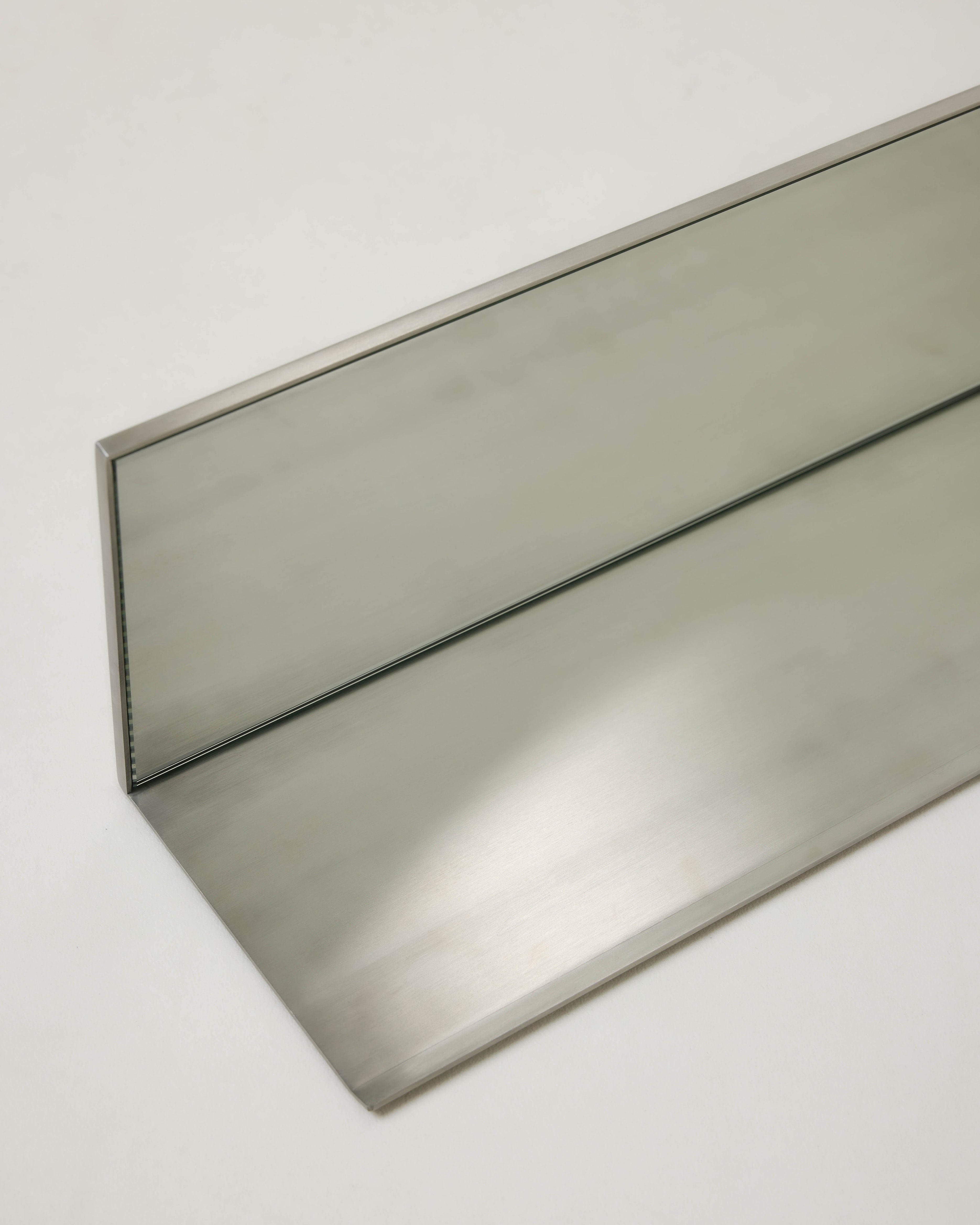 The Mirror Steel Shelf by Dot Objects features a minimalist brushed metal design with a bent edge and central fold, offering a sleek and customizable shelf solution for modern spaces.