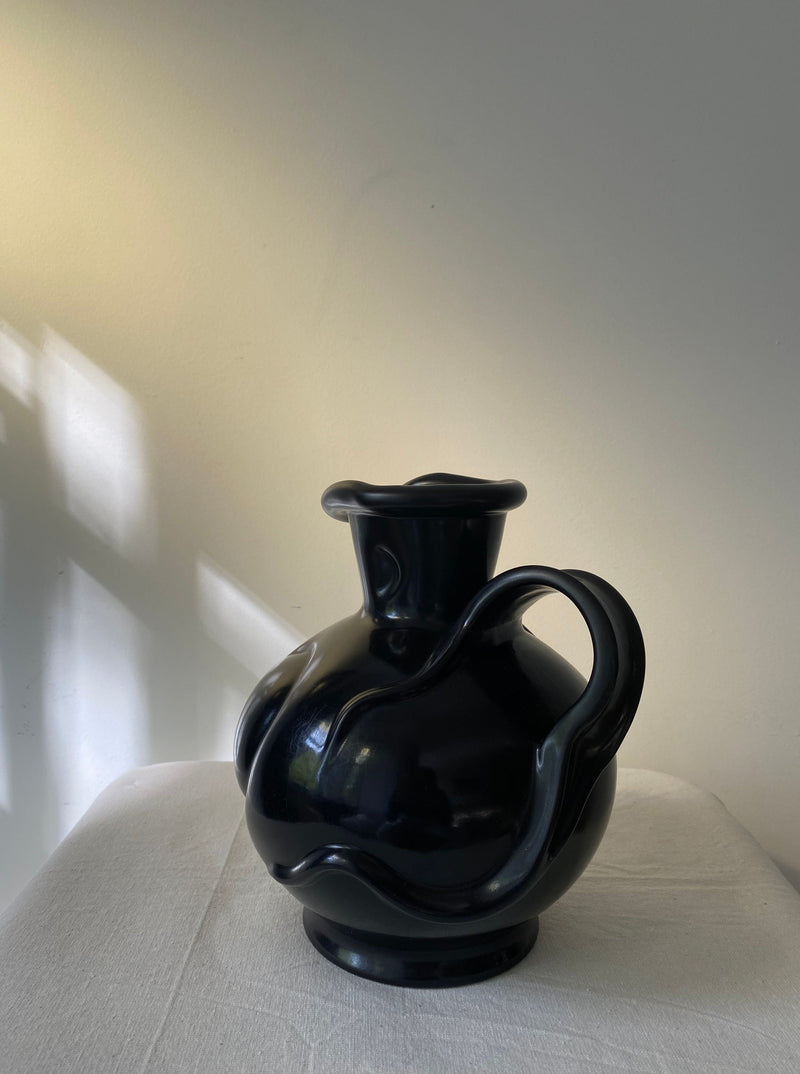 The Art Deco Vase by Les Objets Amsterdam, featuring a glossy black ceramic finish and distinctive wavy handle, rests on a white cloth in soft natural light against a plain pale wall.