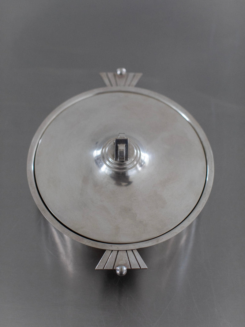 The Out For Lunch Art Deco Bowl is a round, silver bowl with a lid, featuring a geometric handle and decorative triangular and spherical details on opposite sides, reflecting vintage glamour on a grey surface.