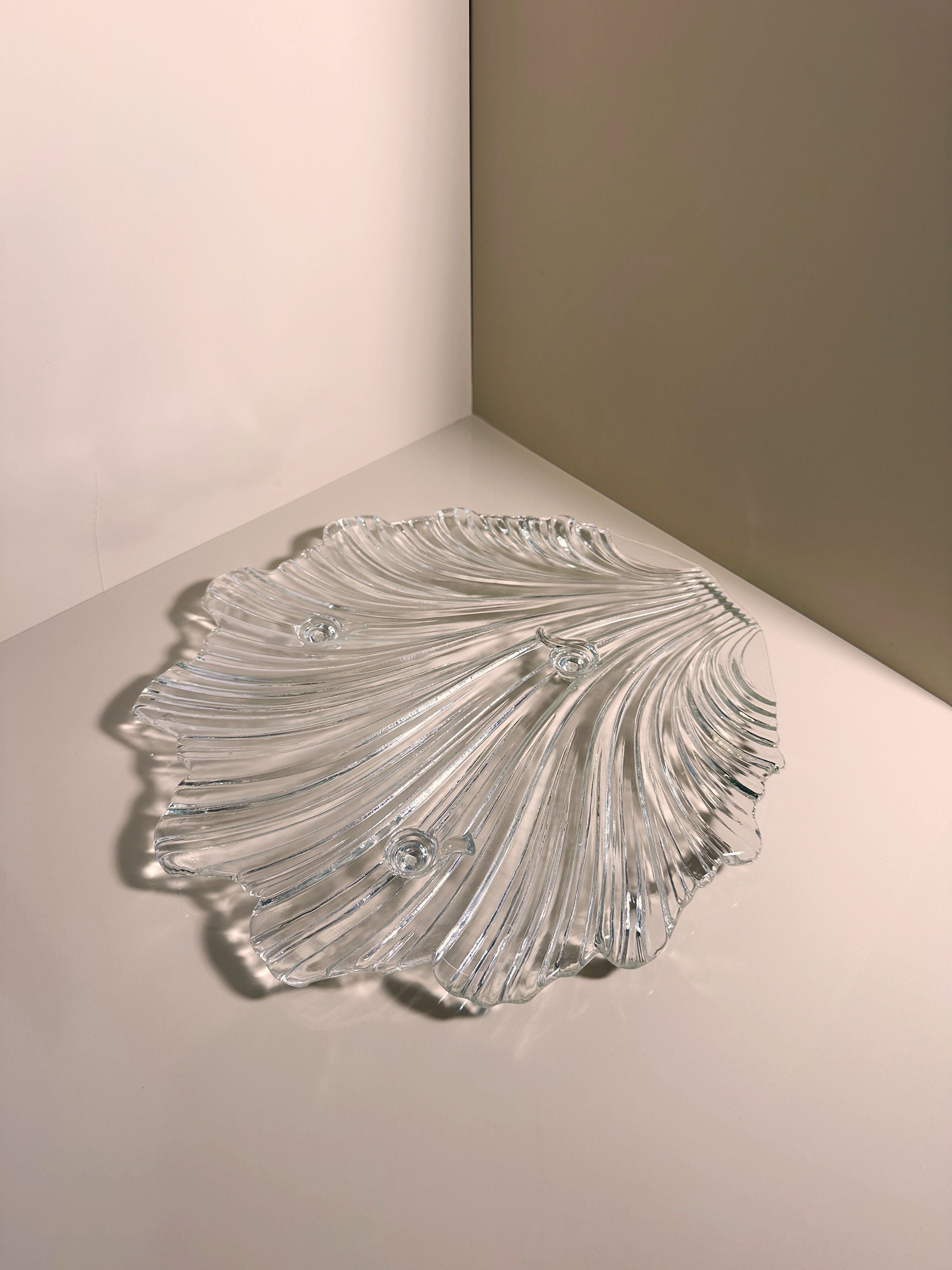The 1980s Shell Shaped Glass Bowl by Dodo Vintage—a clear, wavy glass piece—rests on a white surface, reflecting light and casting delicate shadows for an elegant table setting.