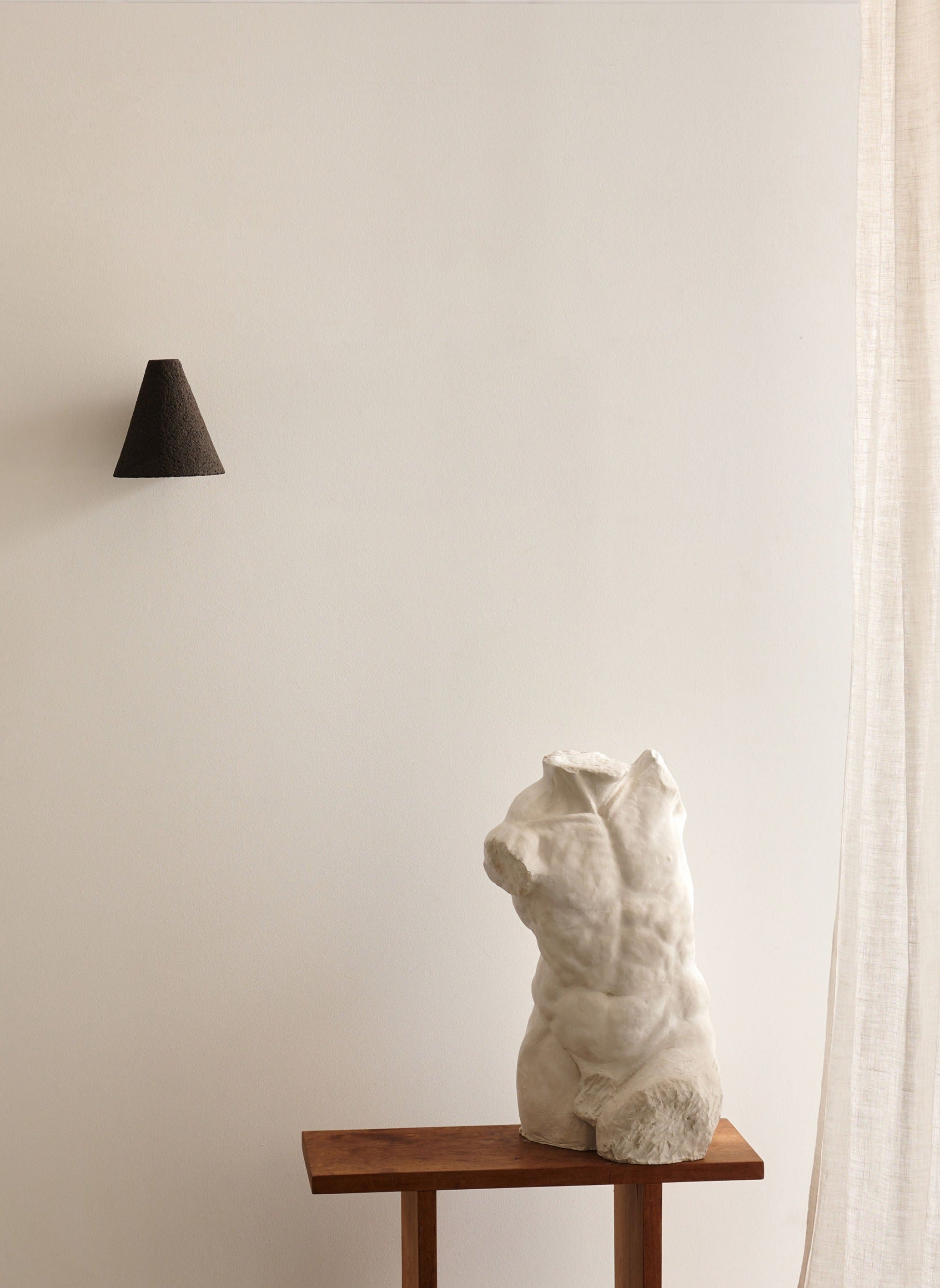 A white classical torso sculpture sits on a wooden table against a beige wall. On the left is the Esoteric Granular Wall Lamp by Mariza Galani, featuring a black cone shape; light beige curtains frame the right edge.