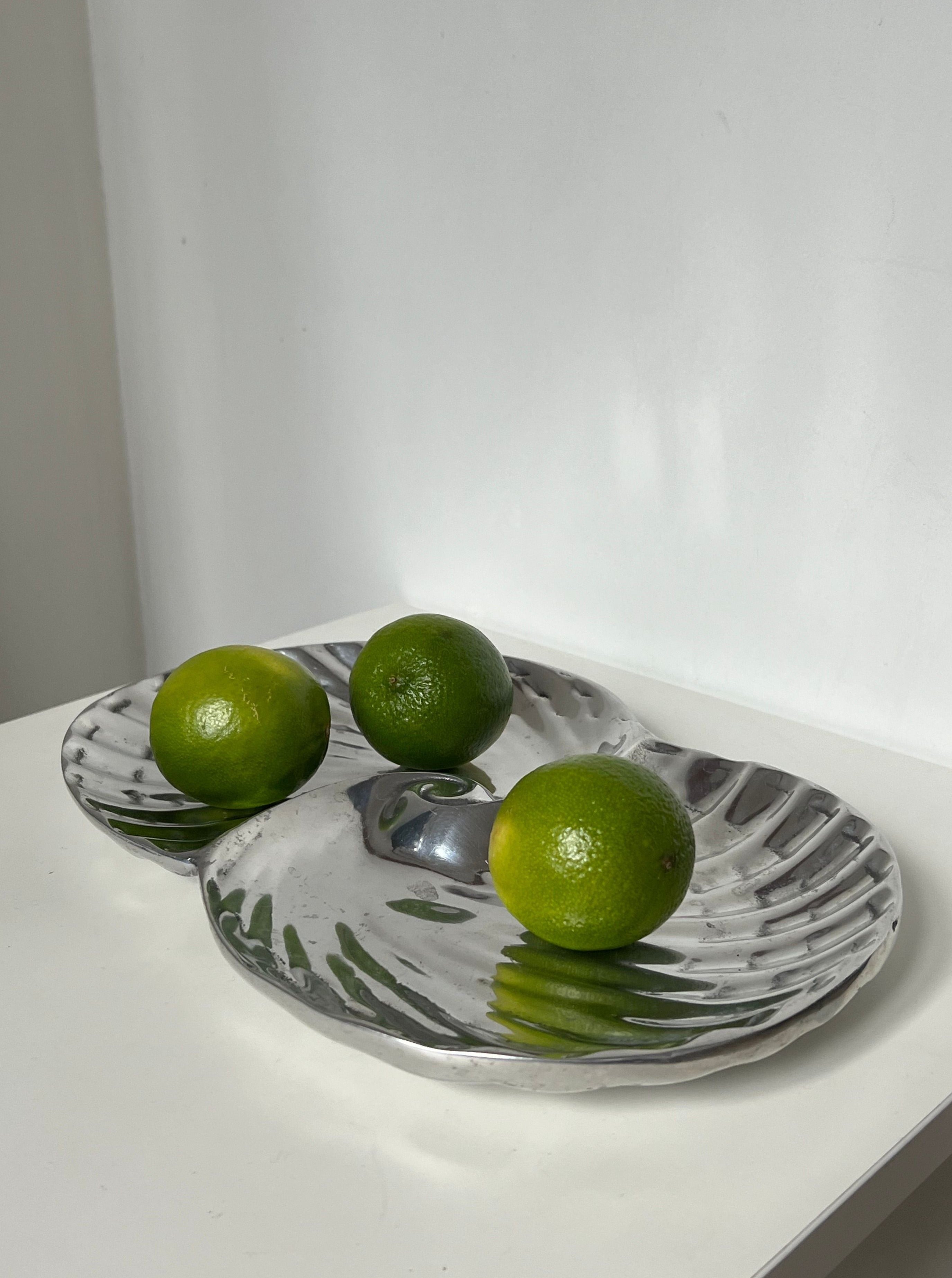 Three green limes rest on the Manushi Swirl Decorative Dish, which shines on a white surface with a plain white wall backdrop.