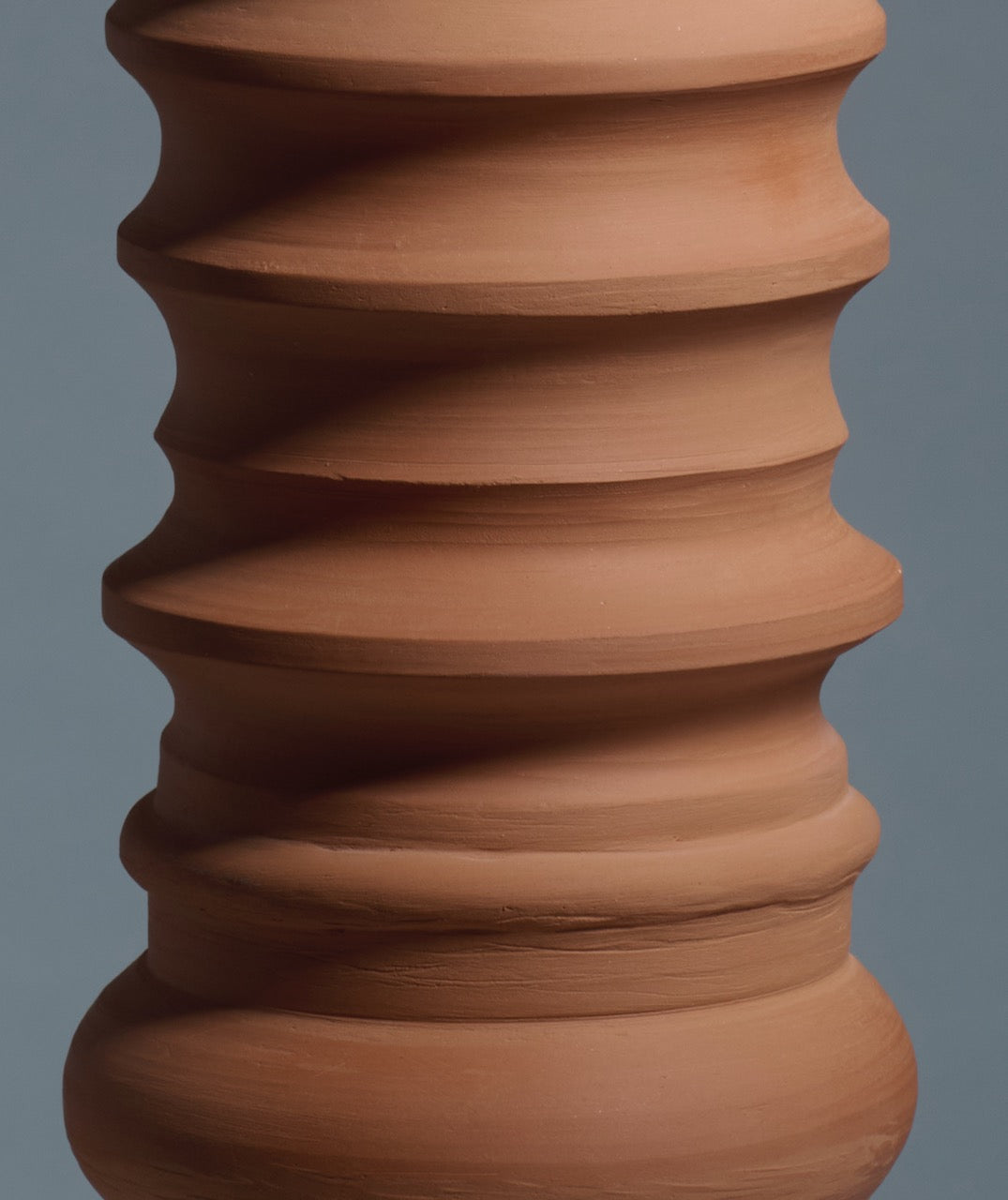 A close-up of Katia Langeard’s Terracotta Glow Table Lamp in Ceramics, featuring ribbed, wavy sculpted grooves and a natural reddish-brown finish against a plain gray background.