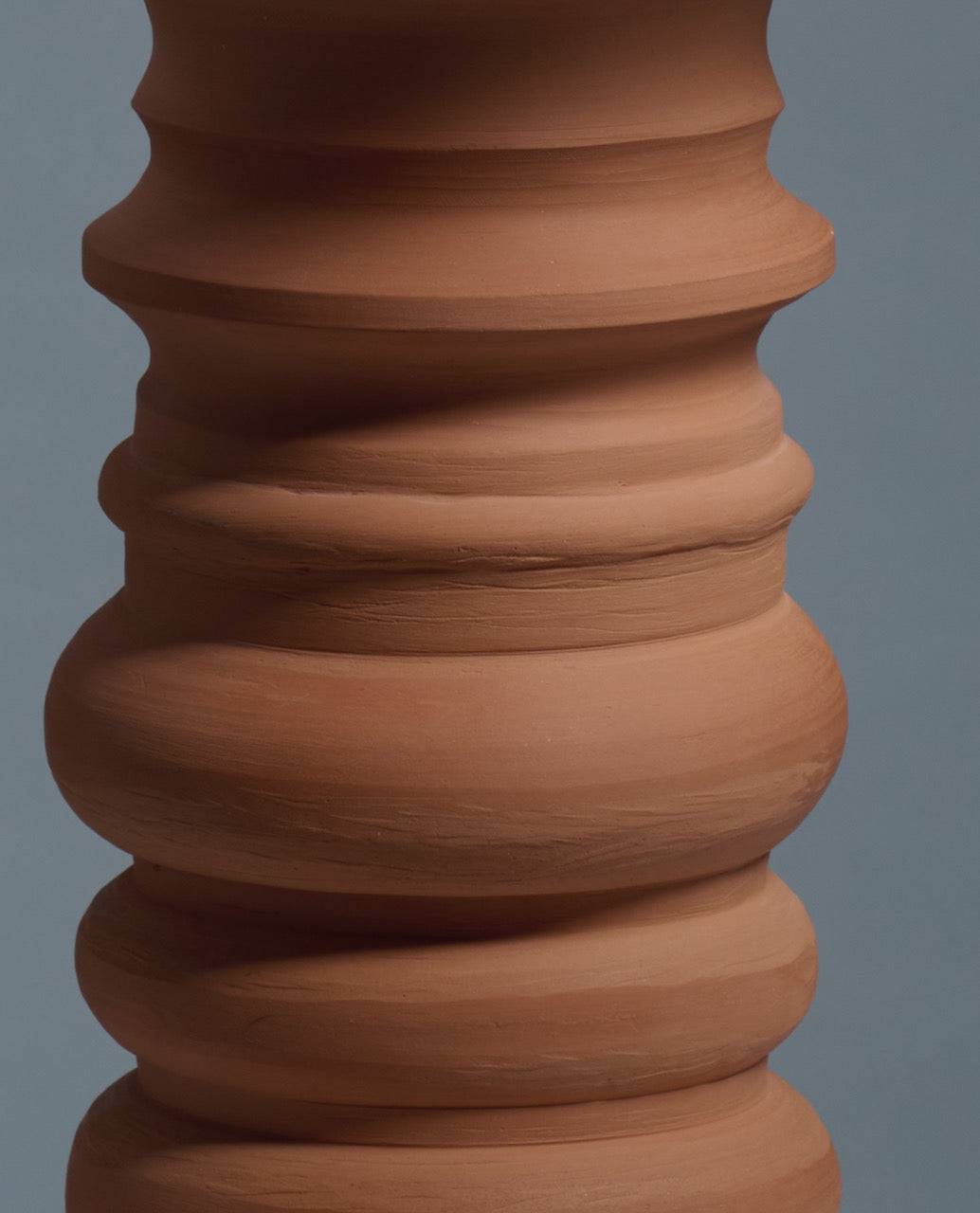 Close-up of a textured, unglazed Terracotta Glow Table Lamp by Katia Langeard, featuring rounded and ridged ceramic forms against a plain blue background.