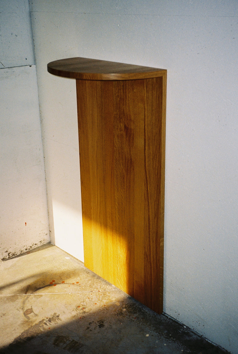The Megaron - Minimalist Shelf in Wood - Natural Oak Large by COSEINCORSO, a handcrafted, semi-circular solid oak shelf with vertical support, is mounted on a white wall above a concrete floor and bathed in sunlight.