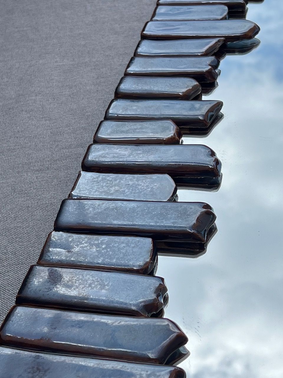 A close-up of Katia Langeard's Cliff Collection Contemporary Mirrors in Ceramic shows dark rectangular tiles lined up by reflective glass, bordered with textured dark fabric, mirroring the sky and clouds.