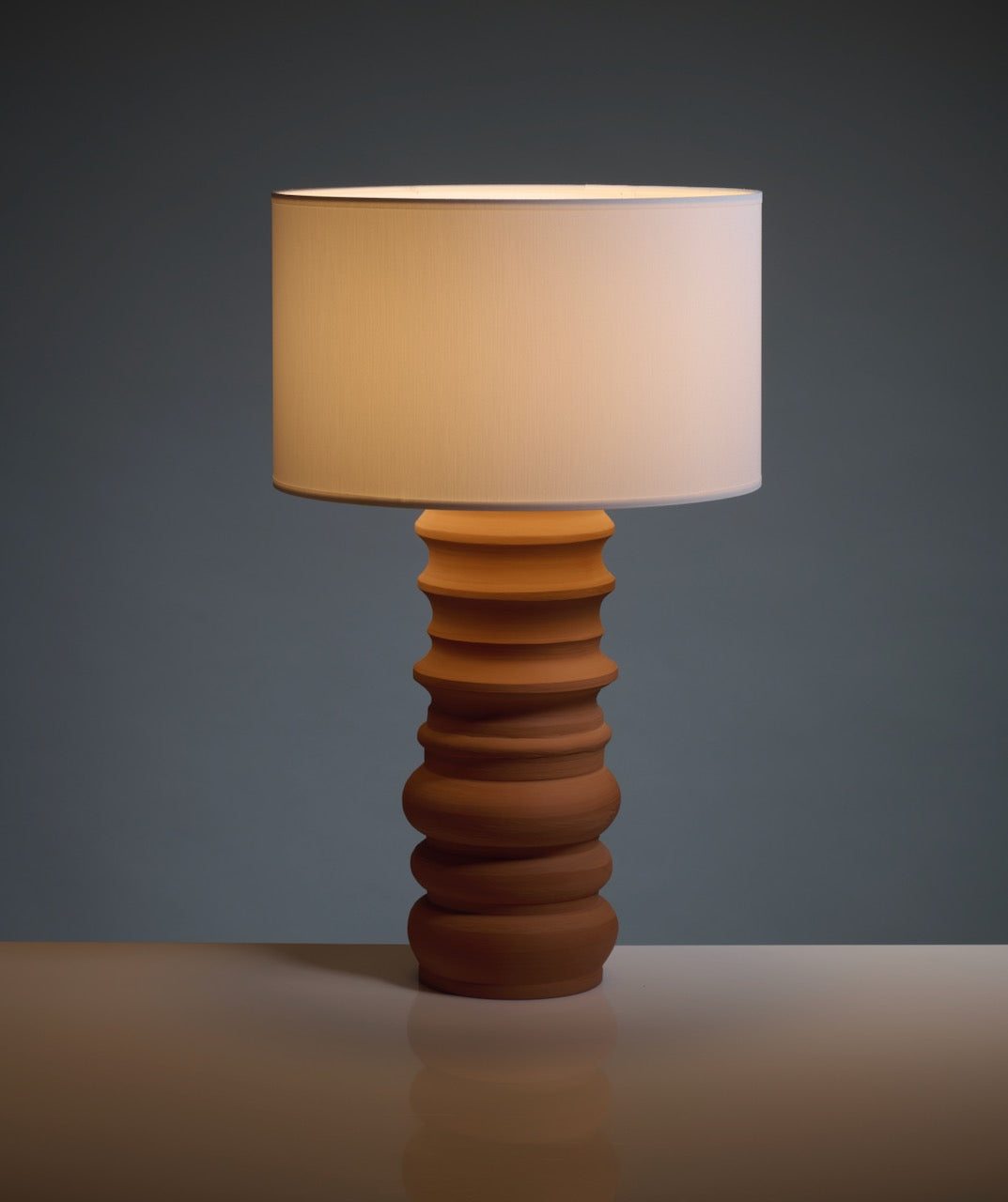 The Terracotta Glow Table Lamp by Katia Langeard features a sculpted ceramic wavy brown base and a large white shade, creating an elegant bedside accent against a dark gray backdrop.