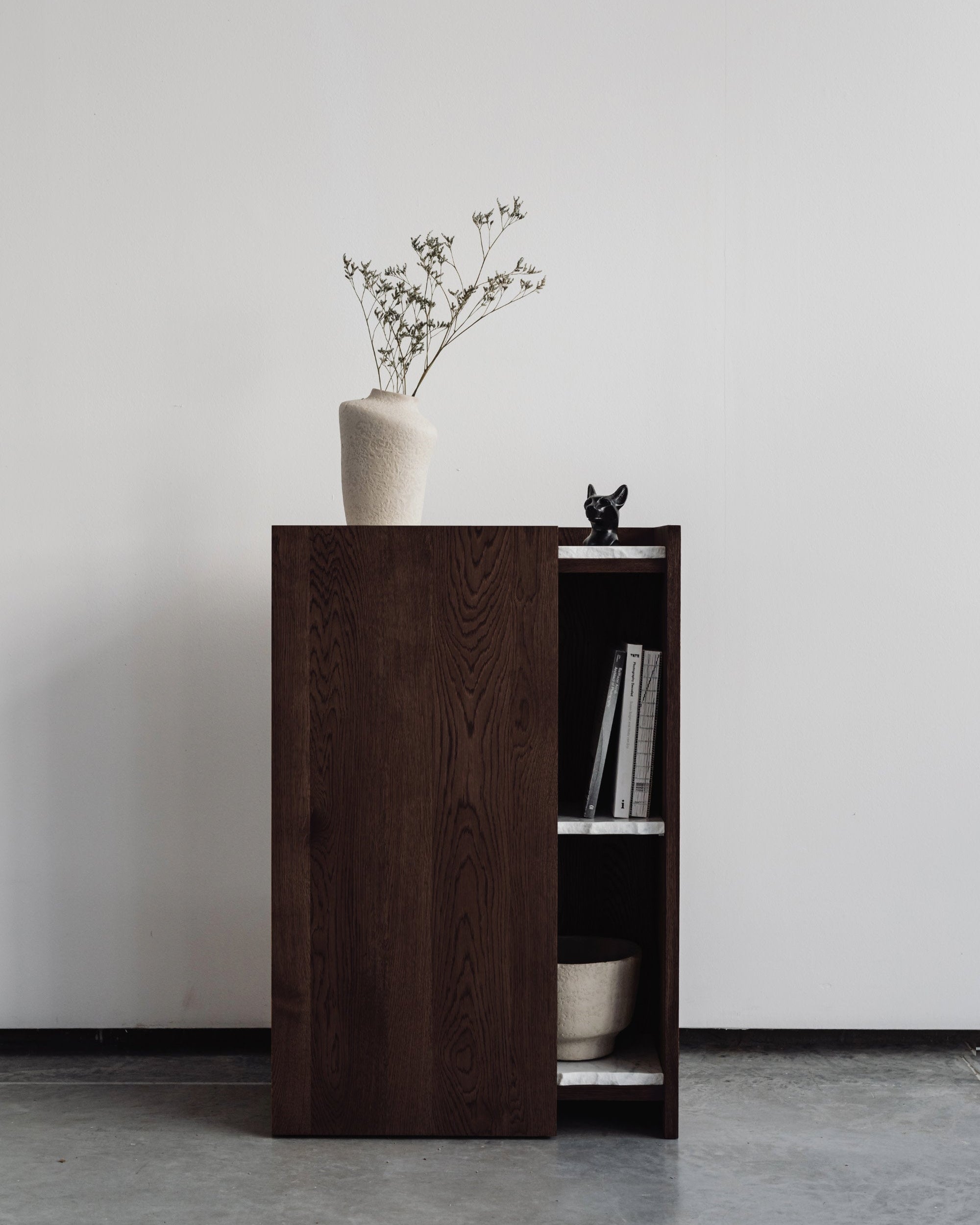 The un'common PAVO Cabinet, a minimalist solid wood storage piece, stands against a plain white wall with a small plant in a white vase and decor on top, while shelves inside hold books and a round beige bowl.