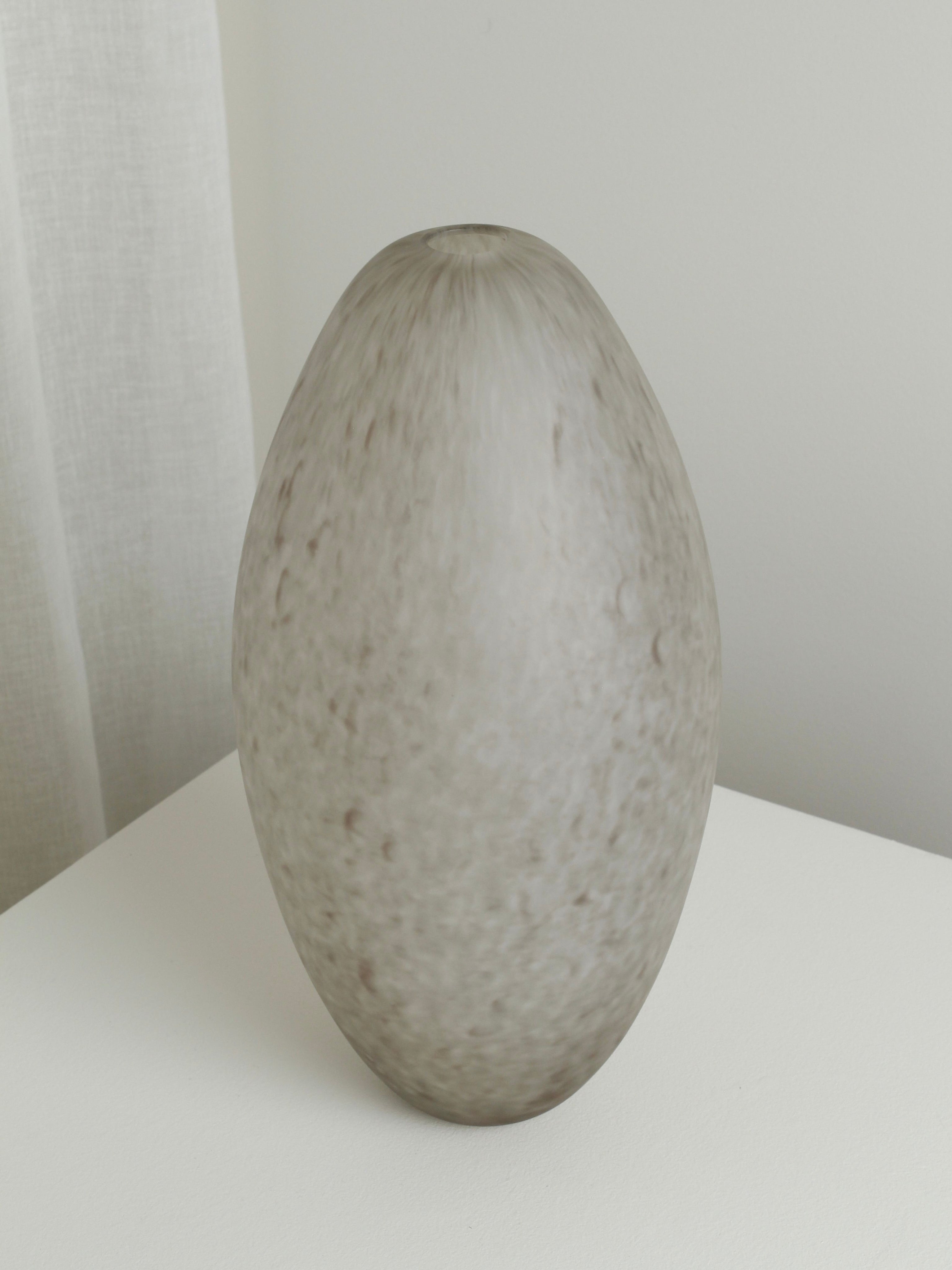 A large, smooth, egg-shaped gray stone with a speckled texture rests on a white surface beside a light curtain and wall, evoking the XLarge Vase in Feathered Taupe by Verre d'Onge.