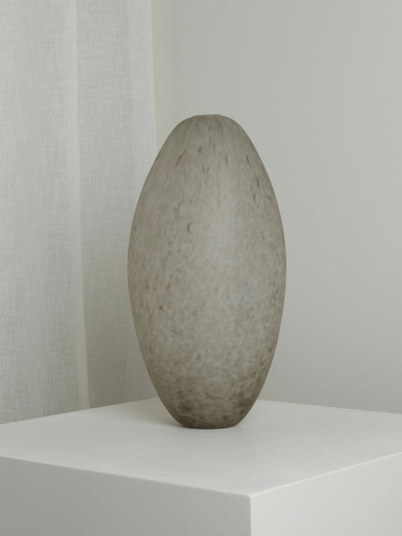 The Verre d'Onge XLarge Vase in Feathered Taupe, with a textured oval shape, sits on a white pedestal against a light gray wall and sheer white curtain.