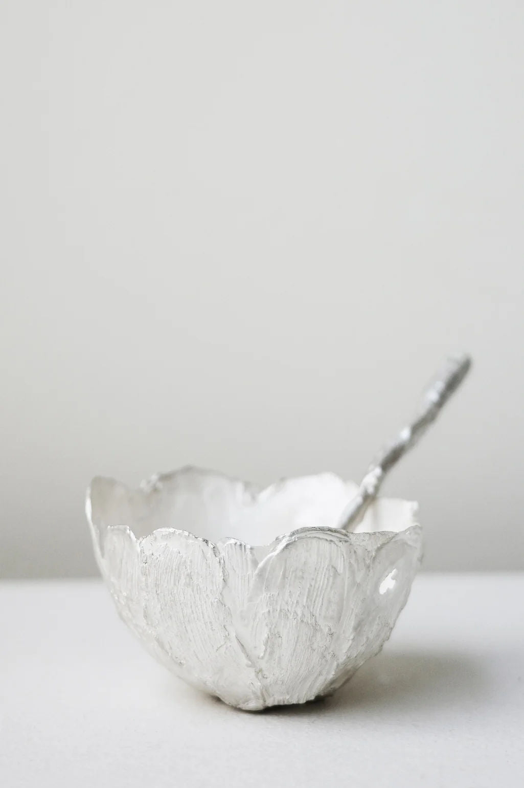 The Completedworks L32 - Spoon in Silver rests in a textured silver bowl with petal-like scalloped edges, showcasing an organic, hand-crafted look against a plain white surface and light background.