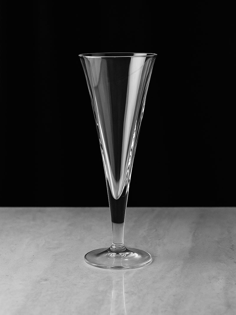 One of six Dodo Vintage Italian Art Deco Revival crystal glasses stands empty on a smooth, light surface against a solid black background, highlighting its tall, conical design.