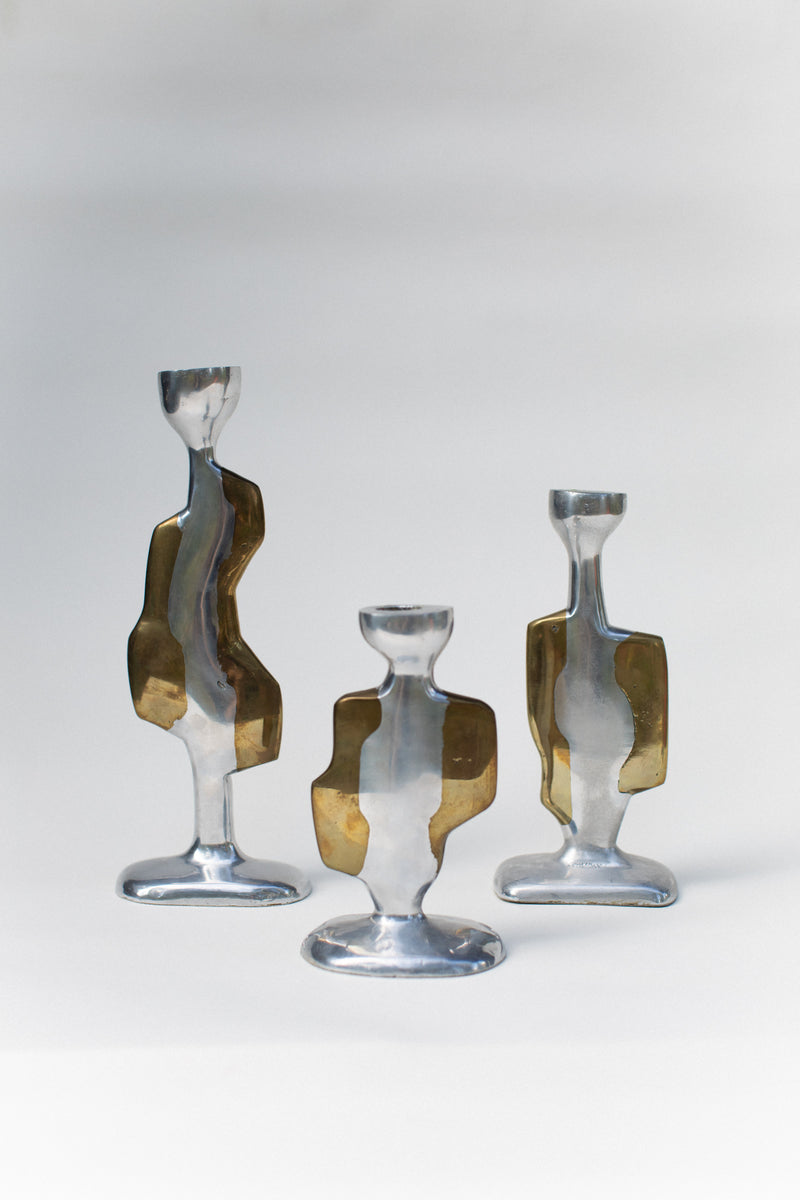 Out For Lunch’s Three Brutalist Candleholders are modern, abstract aluminum pieces with irregular curves and translucent amber accents, displayed on a white surface against a light gray background.