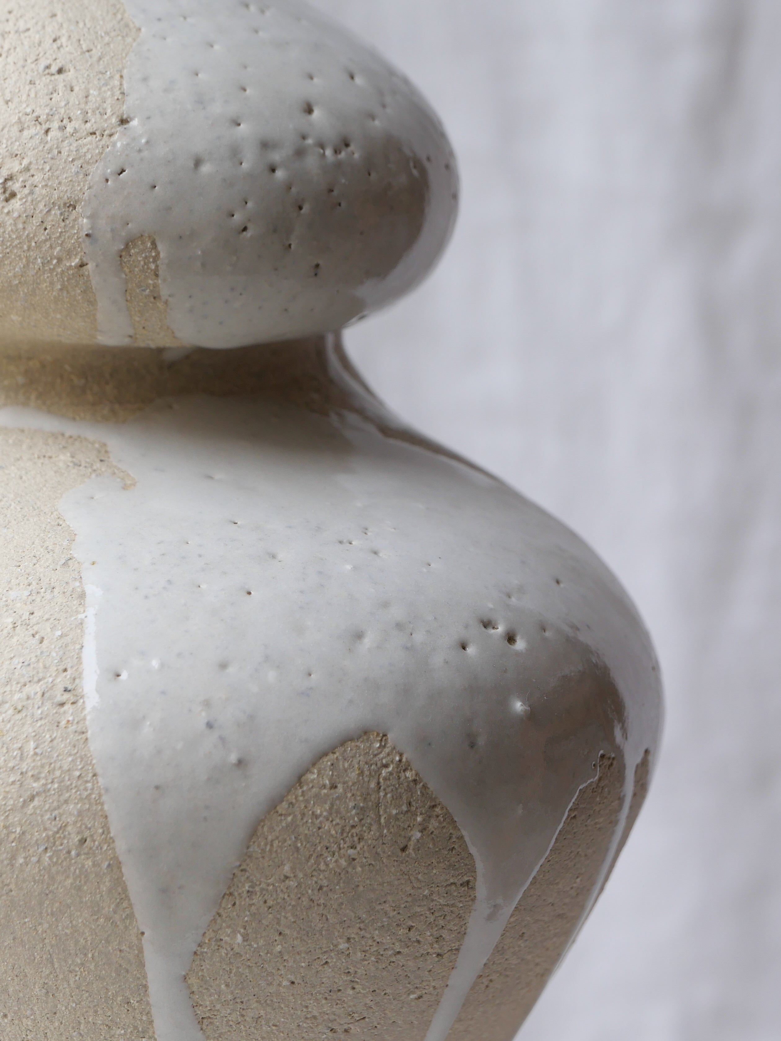 Close-up of the Lucia Mondadori Lilith White Drip: a textured, handcrafted stoneware vase with glossy white glaze flowing down its curved surface, set against a soft, light fabric background.