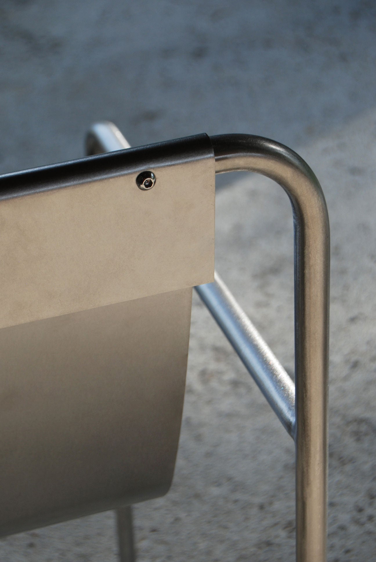 Close-up of SALAK's DESSERT chair, showcasing its minimal design with a brushed steel backrest and tubular frame, set against a textured concrete background.
