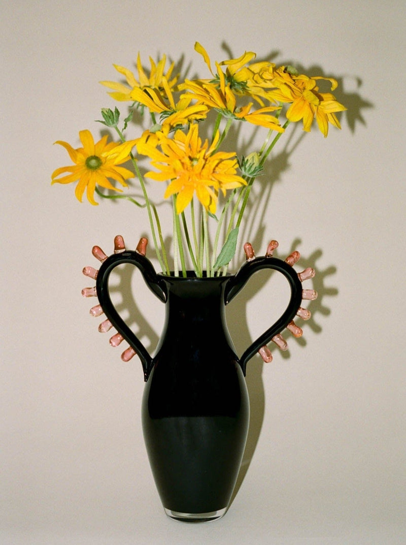 The L'Aurore Vase by Sophie Lou Jacobsen, featuring sculptural handles and decorated with small objects, holds yellow flowers with green stems against a simple backdrop.
