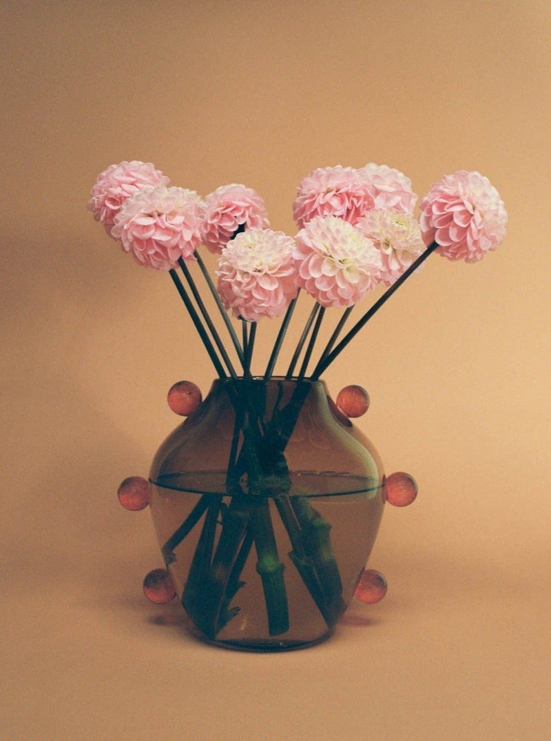 A glass vase with a reflective surface, known as the Sophie Lou Jacobsen La Chaleur Vase, holds a bouquet of pink dahlias, arranged artistically on a warm beige background. The vase is adorned with small